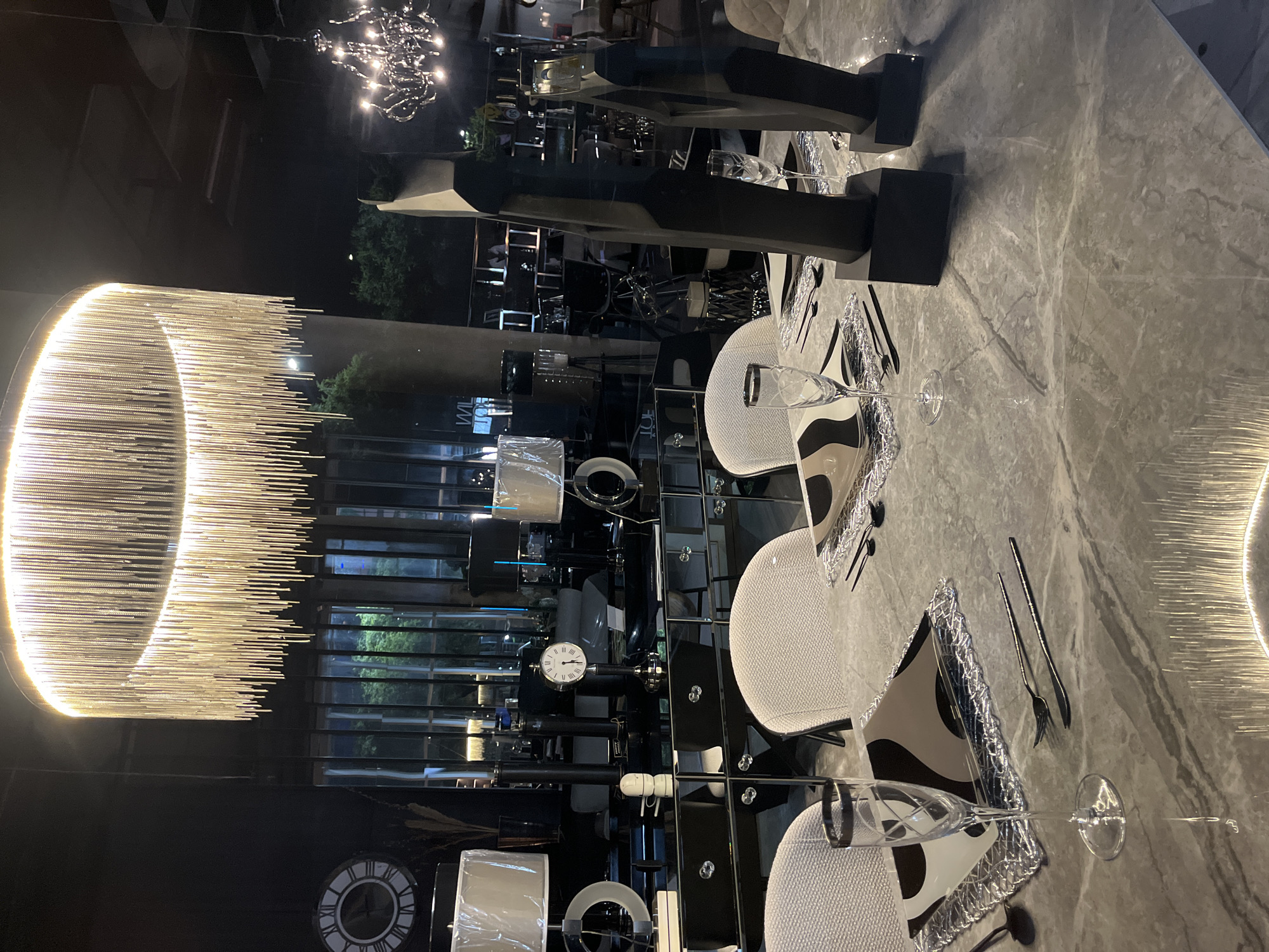 The image presents a high-end interior, seemingly within a furniture store or showroom. Dominating the left side is a large, circular chandelier composed of numerous illuminated rods, positioned against a mirrored wall. This wall reflects the outdoor scene visible through a window, which includes trees and building elements. The foreground features a polished, marble-patterned table meticulously set for a formal dining experience. Place settings are arranged with decorative placemats, elegant cutlery, and delicate glassware. Two chairs with light-colored upholstery are positioned at the table, and abstract, geometric sculptures are placed as centerpieces. A clock and other decorative objects are visible in the reflection on the mirrored wall, suggesting a retail setting. There is no evidence to specify that the image is located in the city of Buenos Aires, Argentina. The image focuses on showcasing high-end interior design and luxury furniture.