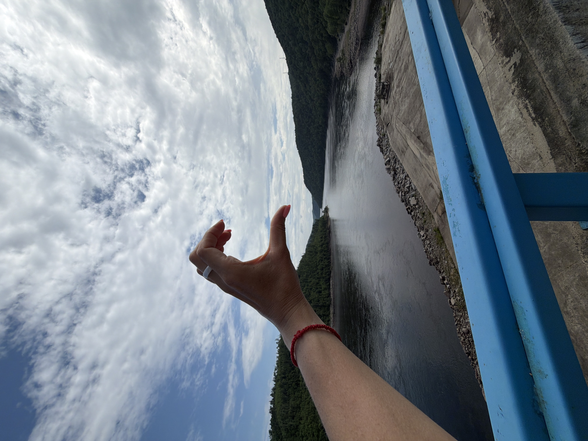A close-up, first-person perspective shot shows a hand with a white ring on the index finger forming a crescent shape against a backdrop of a wide river, lush green hills, and a cloudy sky. The hand, adorned with a red braided bracelet on the wrist, is positioned in the foreground on the left side of the frame. The river occupies the middle ground, flowing between two densely forested hills that recede into the distance. The sky above is filled with a mix of white and gray clouds against a blue expanse. In the lower right corner, a bright blue railing with some signs of wear runs horizontally across the frame, suggesting the vantage point is from a bridge or elevated structure. The location is identified as Talakan, Russia.