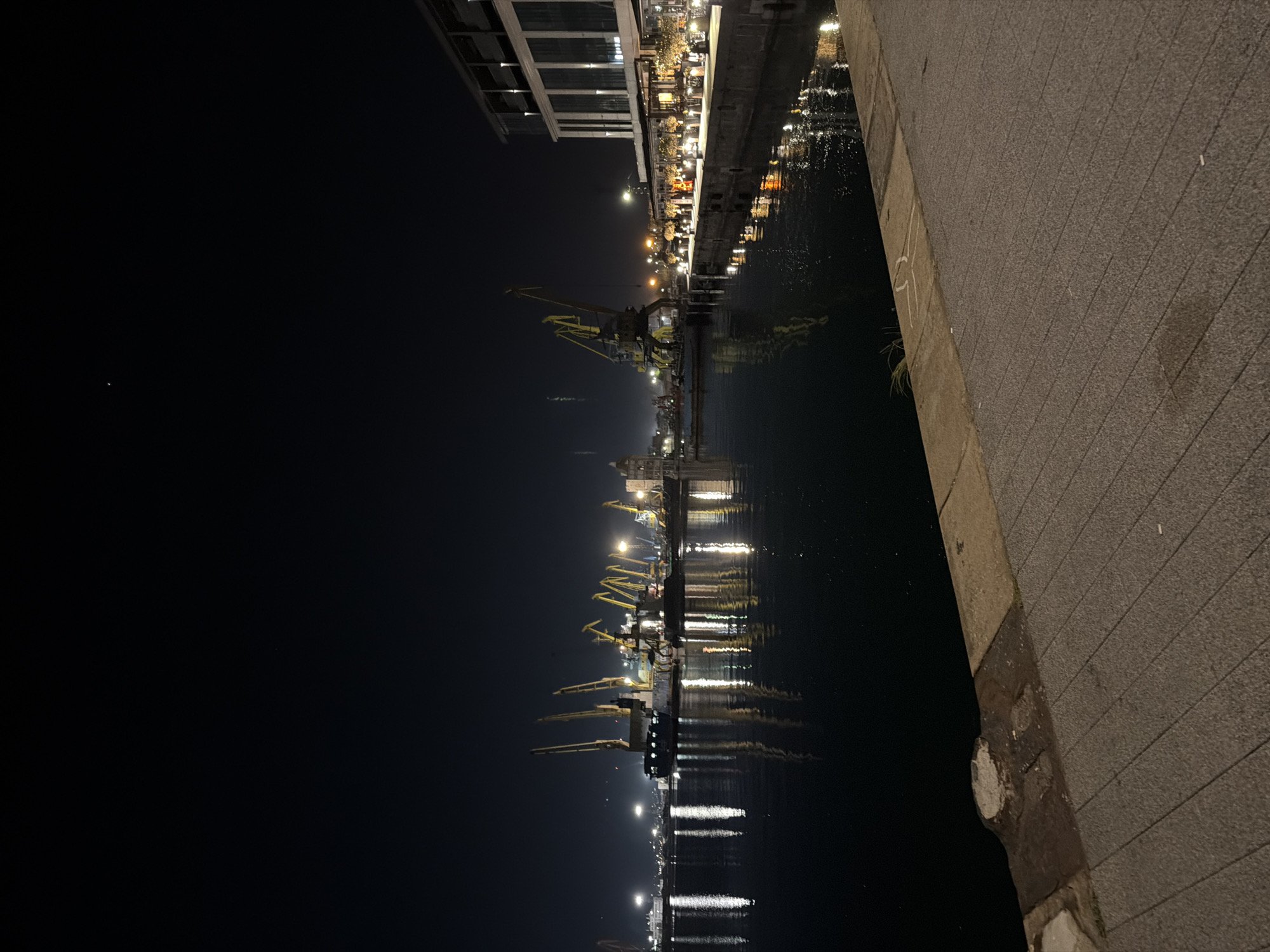 The image depicts a night scene in Burgas, Bulgaria, at the harbor. The foreground shows a paved walkway, with the edge of a quay or pier separating it from the water. Numerous cranes are visible, reflecting in the dark water. On the left, the city of Burgas is mostly dark, but some lights are visible. The upper portion of the image contains a lit building. The image contains no people or interactions.
