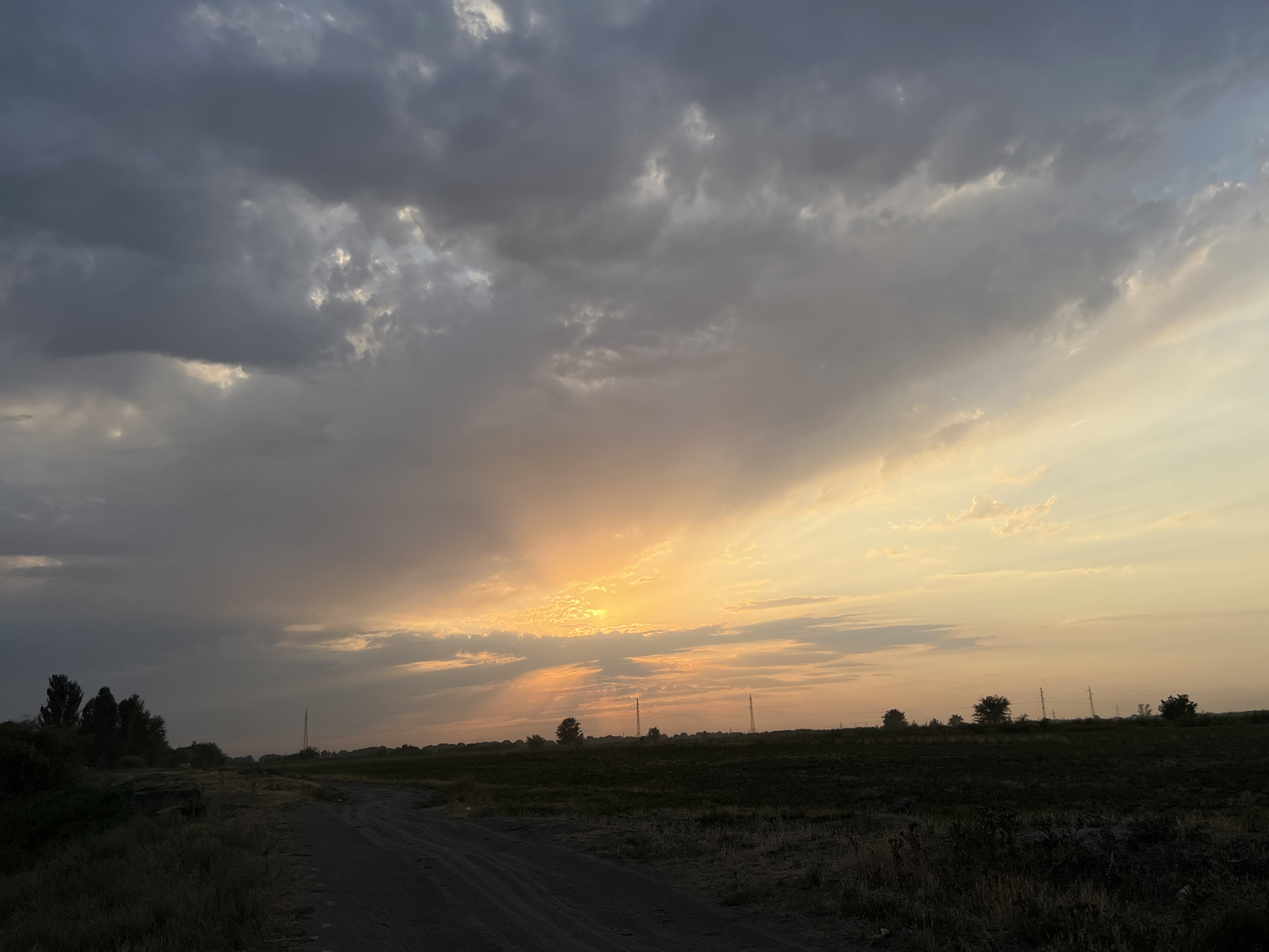 The image depicts a sunset over a rural landscape. In the foreground, a dirt road curves towards the right, bordered by dry grass and vegetation. To the left of the road, a line of dark trees stands against the dusky sky. The middle ground features a flat, dark field, dotted with a few scattered trees and several tall, slender electrical pylons. The horizon is marked by a low, dark silhouette of distant trees and structures. The sky is dominated by dramatic clouds. Dark, heavy clouds occupy the upper left portion of the frame, contrasting with brighter, more diffused clouds towards the right, where the sun is setting. The sunlight breaks through the clouds, creating beams of orange and yellow light that illuminate the sky and cast a warm glow over the scene. The overall impression is one of a serene, yet slightly somber, rural sunset. The location is identified as the city of Drujba, Kyrgyzstan.