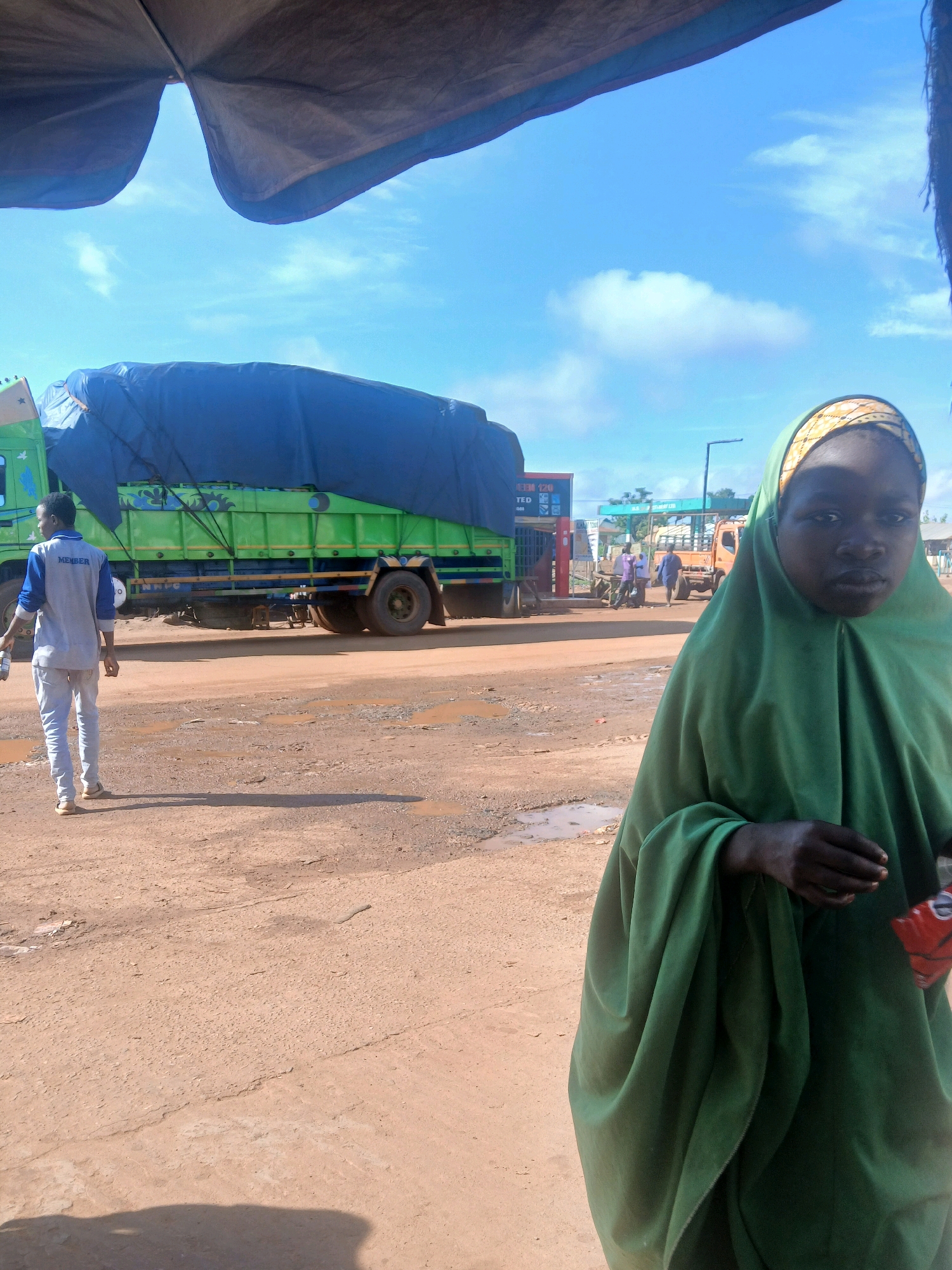 The image depicts a scene in Mokwa, Nigeria, under a bright, cloudy sky. In the foreground, a young girl wearing a green hijab and traditional clothing is looking towards the camera. To the left, a man wearing a grey shirt with "MEMBER" printed on it and white pants stands with his back to the camera, holding a bottle. Behind him, a large green truck covered with a dark blue tarp is parked. Further in the background, there are more vehicles, including an orange truck, and what appears to be a gas station. The ground is a reddish-brown dirt, with puddles of water scattered around, indicating a recent rainfall. The overall atmosphere suggests a roadside setting, possibly a truck stop or market area.