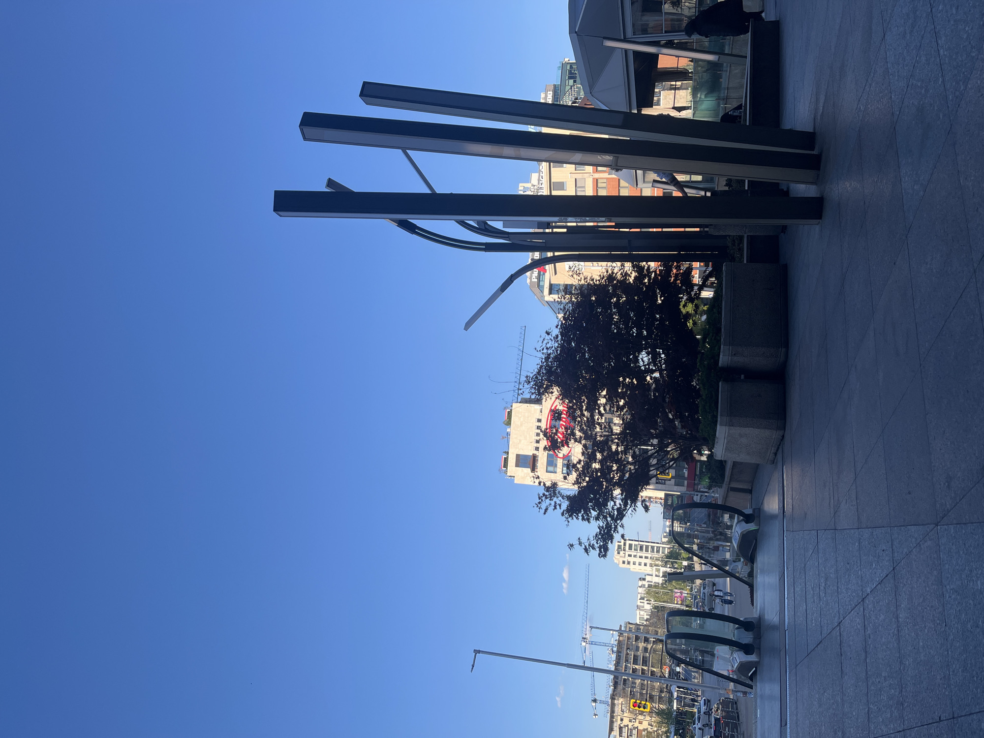 This photo depicts a street scene in Sofia, Bulgaria, taken from a slightly elevated perspective.
In the foreground, there is a gray tiled surface, likely a pedestrian area. Structures resembling modern light fixtures or architectural elements are visible. A small tree with dark leaves sits in a concrete planter.
Beyond this immediate foreground, the view opens to a city street with vehicles, street lights, and buildings. Construction cranes are visible in the distance. The sky is clear and blue.
The architectural style of the buildings in the background appears to be a mix of modern and older structures. The buildings are multi-story. A pedestrian is just visible entering what appears to be an underground transport entrance. The image appears to have been taken during daylight hours.