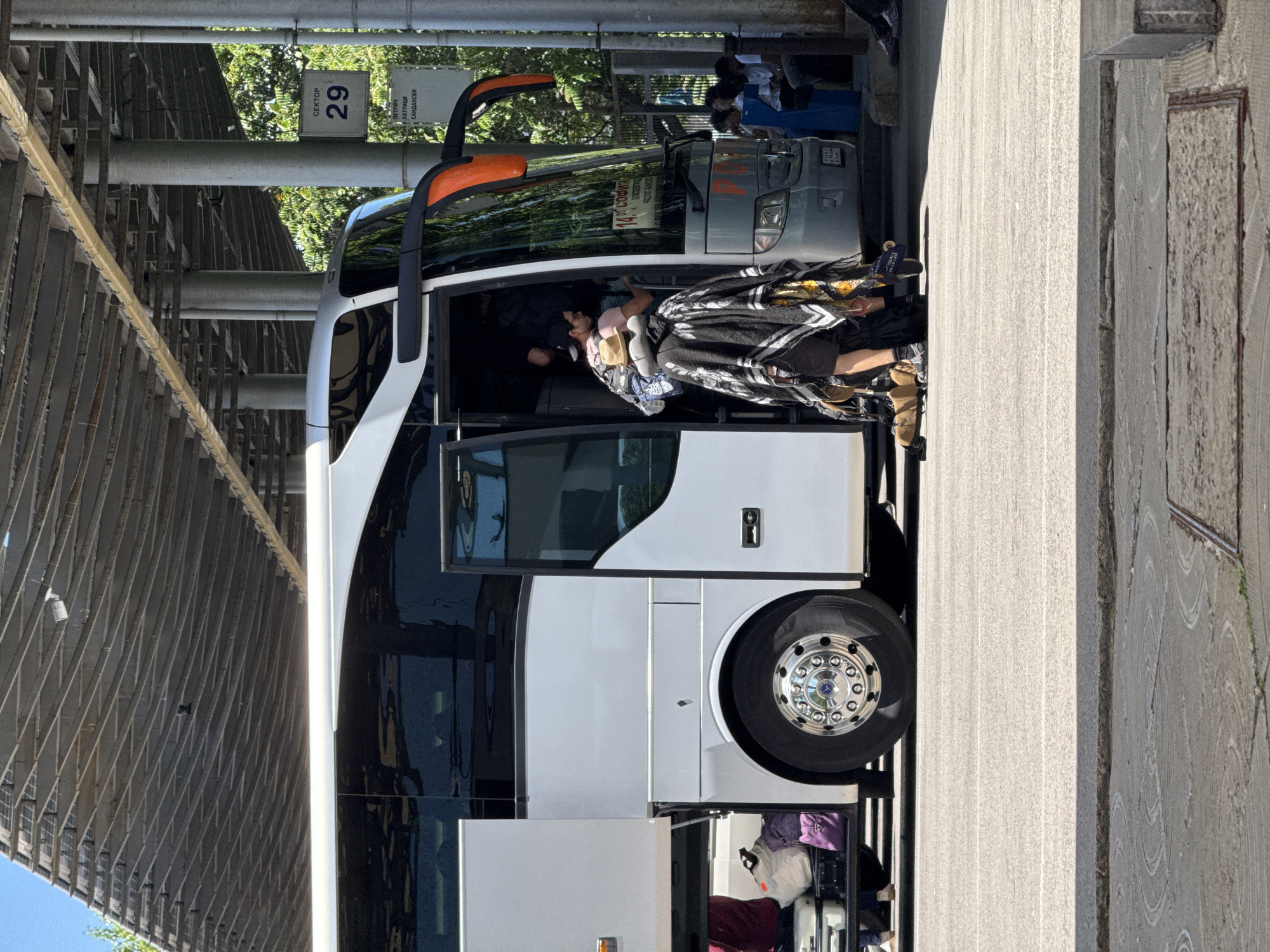 A bus is parked at a bus station with its door open, and people are boarding or disembarking. Luggage is visible in the luggage compartment of the bus. In the background, there is a sign indicating "SECTOR 29" and listing destinations including "PETRICH, KATUNTSi, SANDANSKI". This suggests the location is a bus station in Sofia, Bulgaria, likely serving routes to these southern Bulgarian cities. The bus itself is white with orange accents around the entrance and on the top. The ground is paved, and there is a metal canopy structure overhead supported by pillars. The lighting indicates it is daytime, likely sunny. One person is prominently featured wearing a dark, patterned poncho or blanket over their clothing, facing away from the camera and towards the bus entrance.