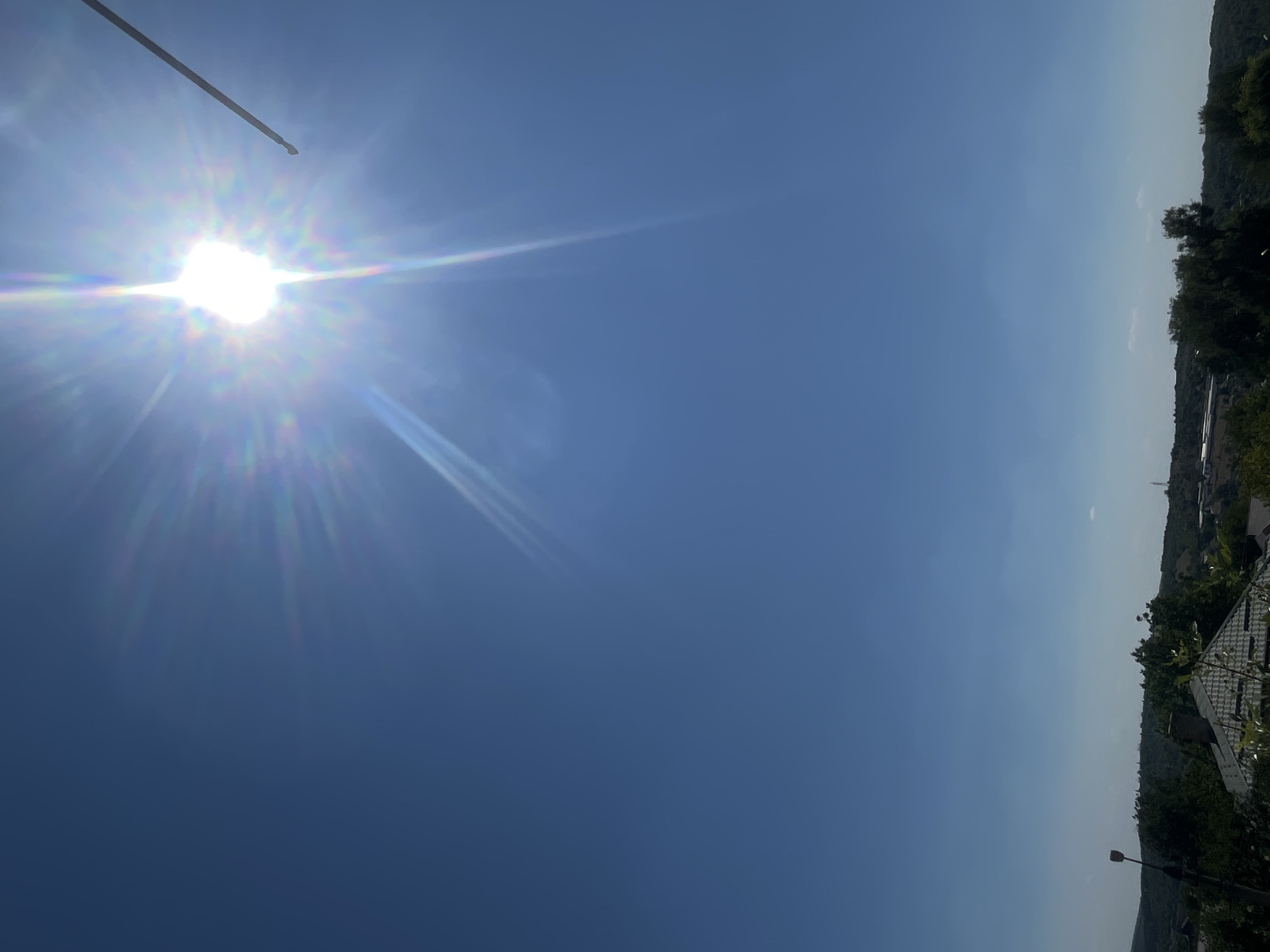 A bright sun with visible rays and lens flare dominates the upper portion of the image, set against a clear blue sky. A thin, dark, linear object extends from the top right corner into the frame. The lower portion of the image shows a landscape with a sloped, tiled roof of a building in the foreground, partially obscured by foliage. Beyond the building, a line of trees forms a treeline against a hilly background. The overall scene suggests an outdoor environment during daylight. The specific location within Rybnitsa, Moldova, cannot be determined from the visual information provided.
