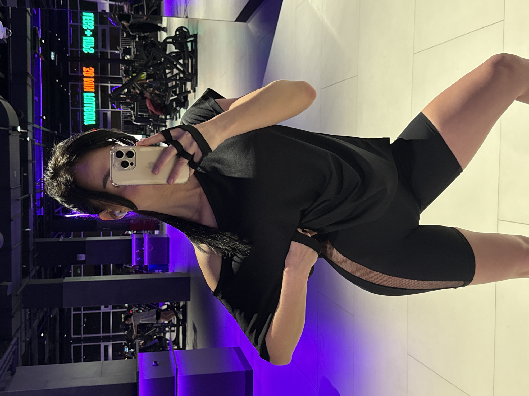 A person, likely female, is captured in a mirror selfie taken within a modern gym or fitness center. The individual holds a light-colored smartphone with a triple-lens camera array, partially obscuring their face. They are dressed in black athletic wear: a loose-fitting t-shirt with cutouts on the shoulders and cycling shorts or leggings featuring a translucent mesh panel along the outer thigh. Fingerless hand wraps are visible on the hand holding the phone.
The background, primarily a reflection in a large mirror, shows various dark, blurred gym equipment. The facility is brightly lit by artificial light, accented by vibrant purple and green neon signs. One prominent neon sign, reflected, reads "30 MIN ЦЕЛЕТВОЙ" (likely intended as "30 MIN TARGETED") and "ТЕСЛ + HIIЭ" (suggesting "TESL + HIIT," possibly referring to a High-Intensity Interval Training program or brand). The floor is light-colored and reflective, contributing to the sleek, contemporary aesthetic. The setting appears to be an indoor gym, with the artificial lighting indicating it could be evening or night. The image captures a moment of self-documentation within a dedicated fitness environment.