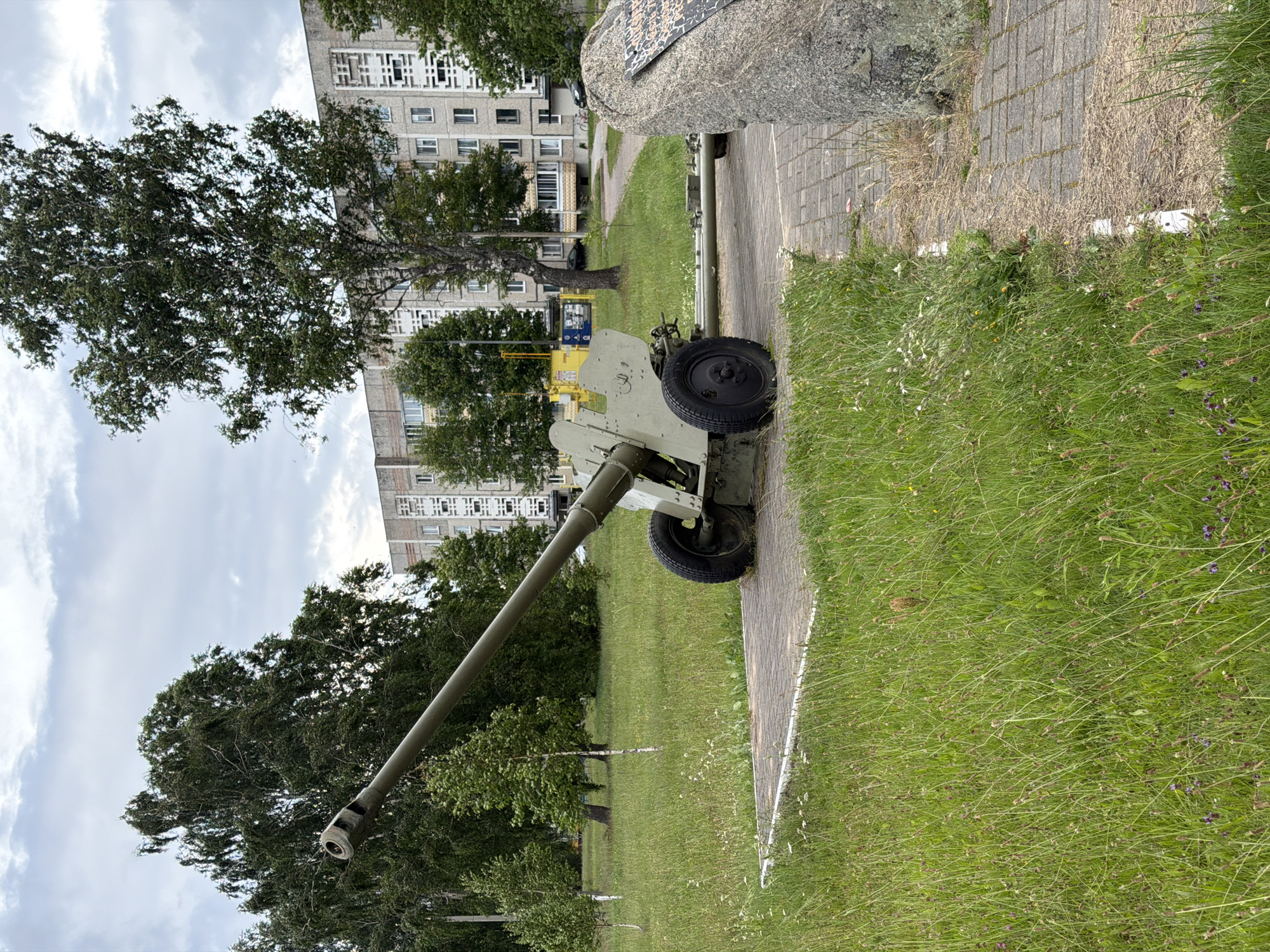 A static, green-painted artillery piece, likely a howitzer, is displayed on a concrete platform surrounded by grass and wild flowers. The gun is positioned at an upward angle, with its barrel pointing towards the left of the frame. It has two large wheels and a trailing mechanism. Behind the gun, a grey, multi-story apartment building with numerous windows and a distinctive architectural pattern on its facade dominates the background. Several tall trees with dense foliage are scattered throughout the scene, with the largest tree positioned prominently behind the gun and to the right. A large, rough-hewn stone memorial, with an inscription partially visible, stands to the right of the artillery piece. The sky is overcast with grey clouds. The scene is located in an outdoor urban setting, possibly a park or memorial site, within the Бягомльскі сельскі Савет region of Belarus. There are no people visible in the image.