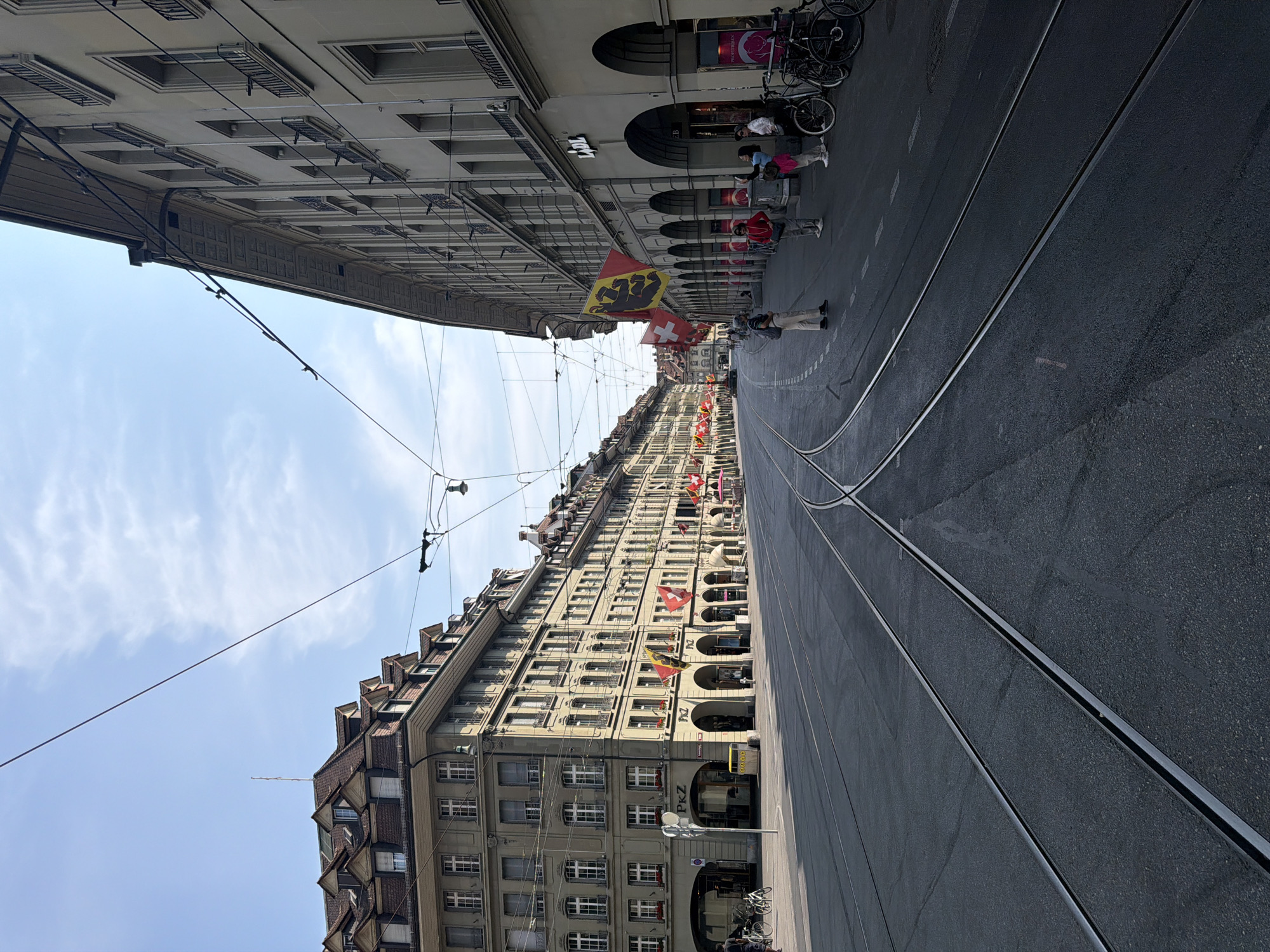 The image depicts a wide, empty street in Bern, Switzerland, with tram tracks dominating the foreground. The street is lined with multi-story buildings featuring arcades on the ground floor. Flags, some appearing to be Swiss flags, are hung from the buildings.
On the left side of the street, a large building with a Mansard roof and numerous windows is visible. A sign that reads "PKZ" is mounted on this building. Across the street, on the right, another long building with arcades is seen. Signs for "Lidl" and "Profi-Boutique" are identifiable on this building.
Several people are present on the sidewalk on the right side of the street. Some are standing, while others appear to be walking or taking photos. Bicycles are parked on the right sidewalk. The sky is partly cloudy, and overhead wires for what are likely trams crisscross the scene. The overall impression is that of a quiet, historic city street in Bern.
