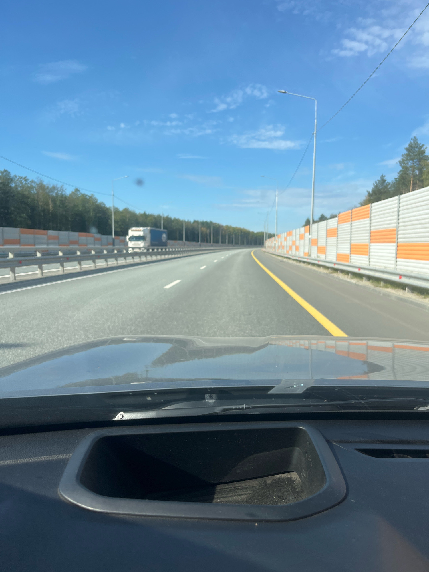 A first-person view from a vehicle traveling on a highway in Bol'shie Algashi, Russia. The road is paved with asphalt and has multiple lanes marked with white lines. A bright yellow line demarcates the edge of the driving lane on the right. On the right side of the highway, there's a sound barrier with orange and white panels, followed by a line of trees. On the left side, a metal guardrail separates the highway from a tree line. A white truck with a blue logo is visible in the distance, traveling in the same direction. The sky is clear blue with scattered white clouds. Streetlights are visible along the highway. The dashboard of the vehicle is in the foreground, showing a dark gray, textured surface with a cubbyhole.