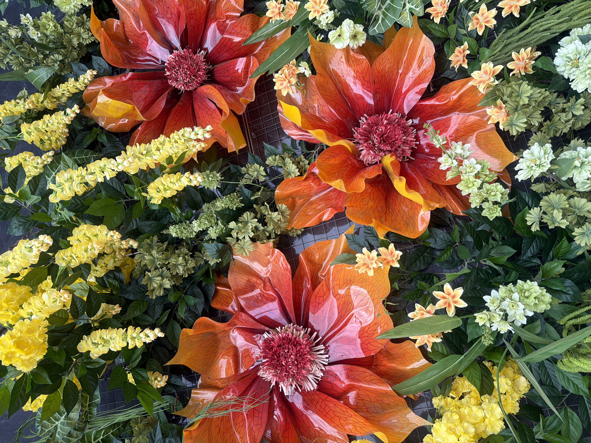 The image depicts an arrangement of flowers. Three large, stylized flowers with orange and red petals are the focal point. Smaller yellow and white flowers are interspersed among the larger blooms, along with green foliage. The scene is likely a decorative display. There is no indication of the location being Rostov-on-Don, Russia, nor any people, subjects, objects, actions, or interactions beyond what is described above.
