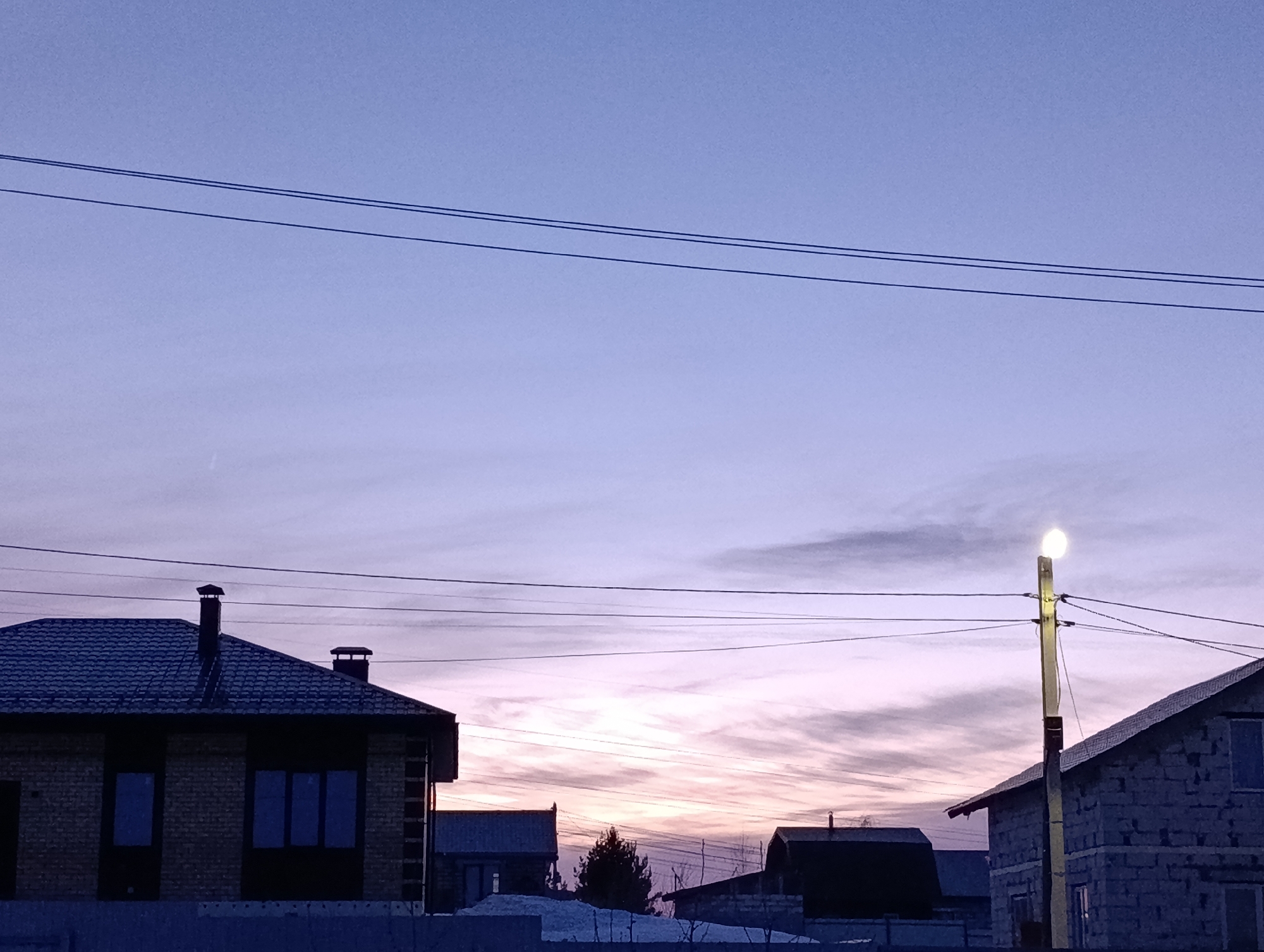 The image presents a residential street scene in Chaikovsky, Russia, at either dawn or dusk, indicated by a sky gradient transitioning from muted blue overhead to pale pink and orange hues near the horizon, populated by wispy clouds. Multiple dark utility lines traverse horizontally across the upper and middle sections of the frame. On the left, a two-story brick house with a dark, multi-panel roof and two dark chimneys is visible, featuring dark-framed windows. To the right, a structure constructed from light-colored masonry blocks, topped with a dark roof, is partially visible. A utility pole with an illuminated street light stands prominently between the two main structures on the right side. In the mid-ground, a dark evergreen tree is discernible amidst other distant house silhouettes. The foreground and visible ground areas are covered with patches of snow. No people or active interactions are present in the scene.