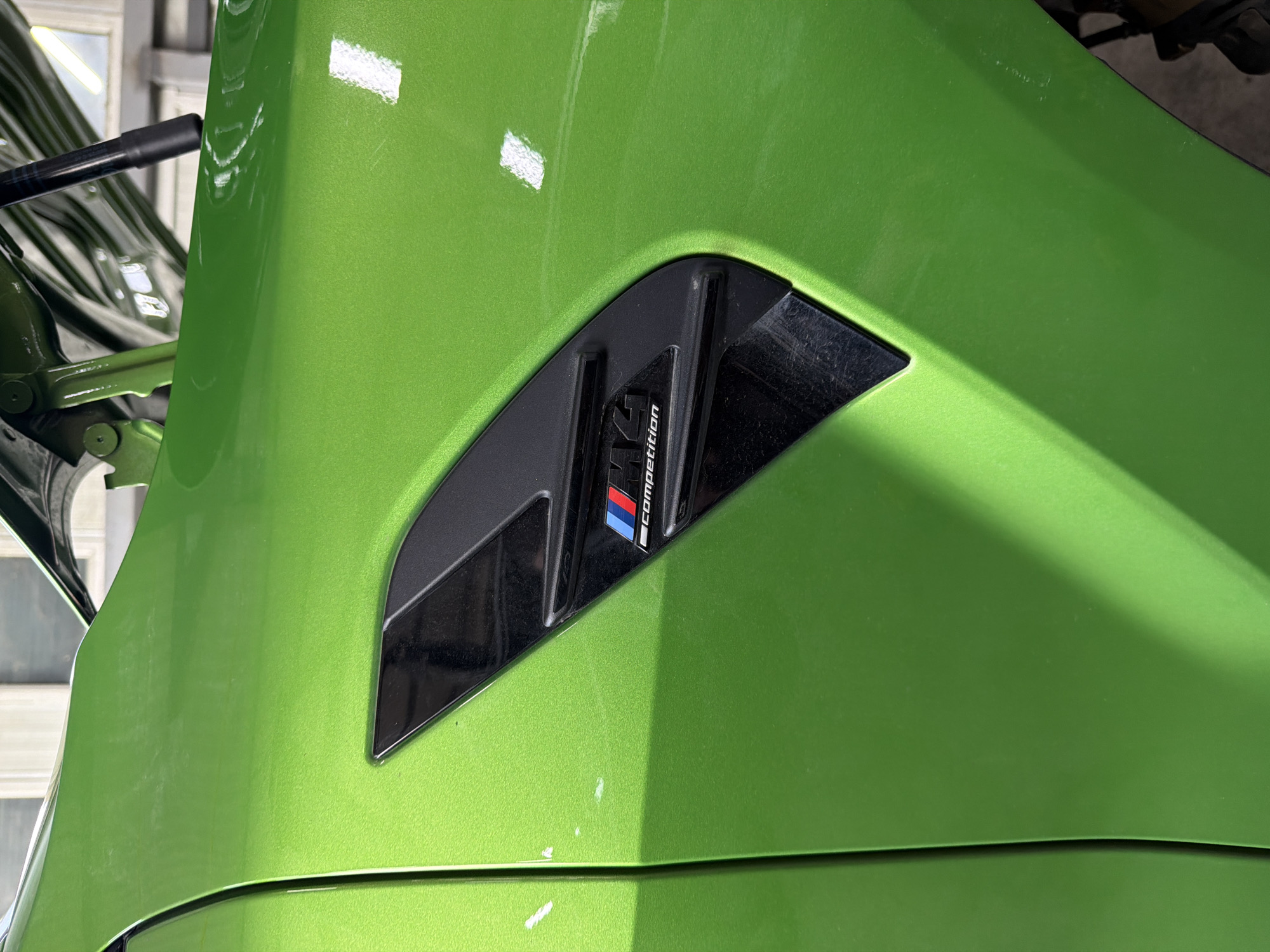 The image shows the green front fender of a BMW M4 Competition. The M4 Competition badge is visible on the fender, located on a black vent. The paint is a bright, glossy green. The angle of the shot suggests a close-up view.