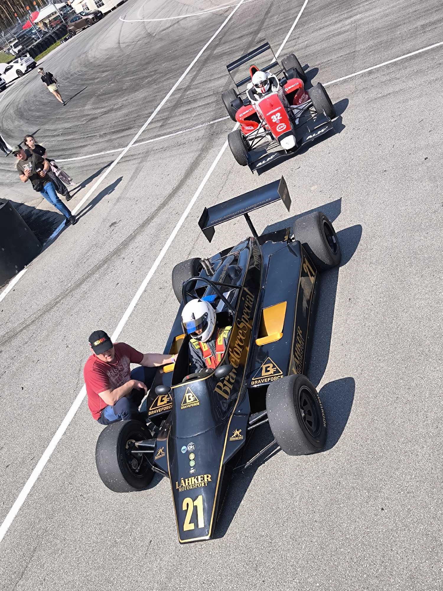 The image shows two open-wheel race cars on a paved track, likely a circuit. The foreground car is predominantly black with gold accents and the number 21 on its front. Markings on the car indicate "Braveforce Special" and "LÄHKER MOTORSPORT". A man in a red shirt and black baseball cap is kneeling near the driver's cockpit of the black car, potentially assisting the driver. The driver is wearing a white helmet with a blue visor. The other car is red with black accents and the number 32 on the front. It is further away from the camera and positioned on the track behind the black car.
In the background, several people are standing or walking on the trackside, and other vehicles are visible in the distance. The track is surrounded by white lines. The location of the photo is at Biķernieki race track, Riga, Latvia.