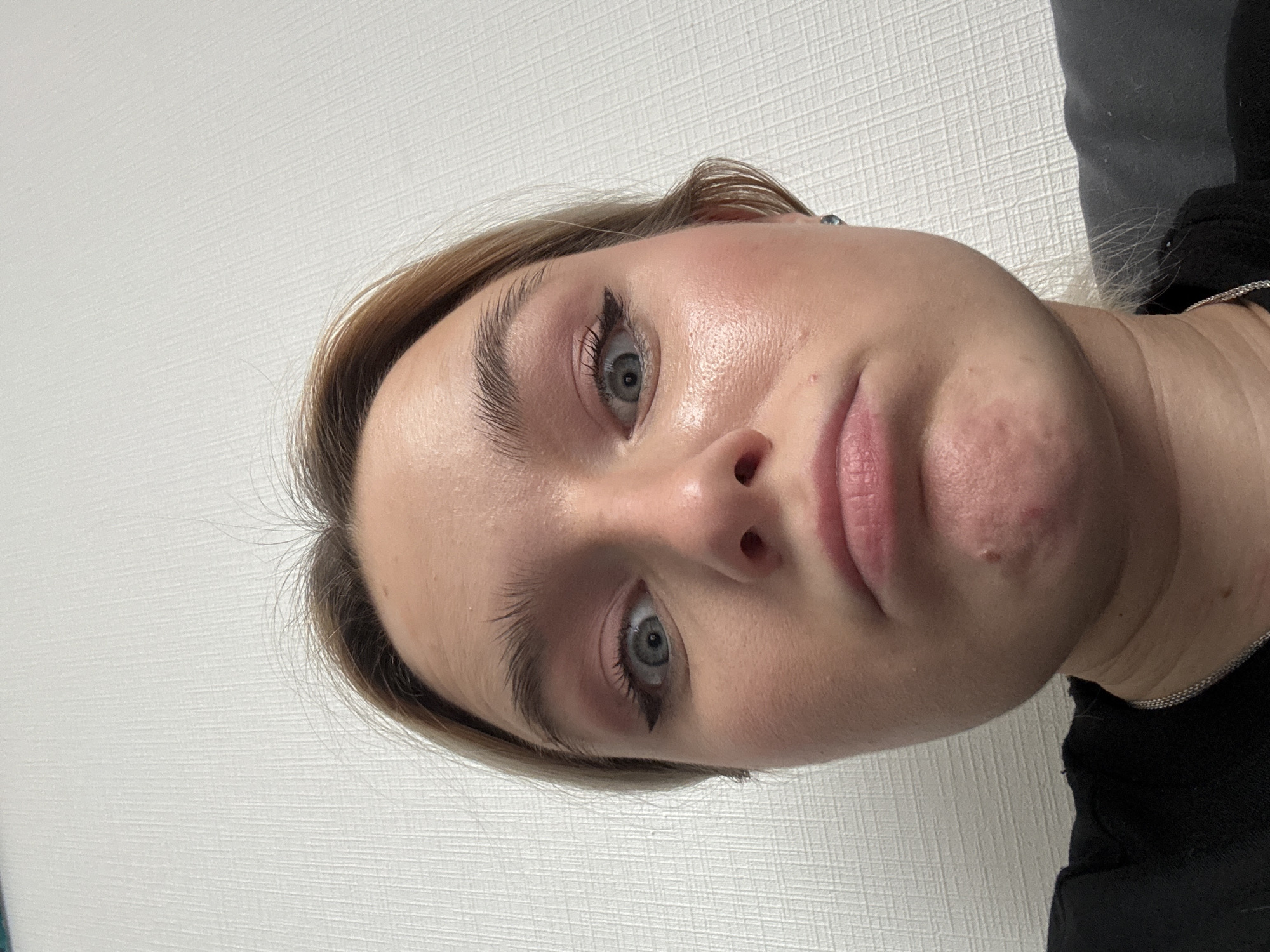 A close-up, tilted shot of a young woman's face. Her skin appears slightly flushed. She has light brown hair, light eyes, and subtle eyeliner. Her lips are pressed together in a neutral expression. The background is a plain, textured white wall on the left side and dark clothing on the right side. The location is unknown.