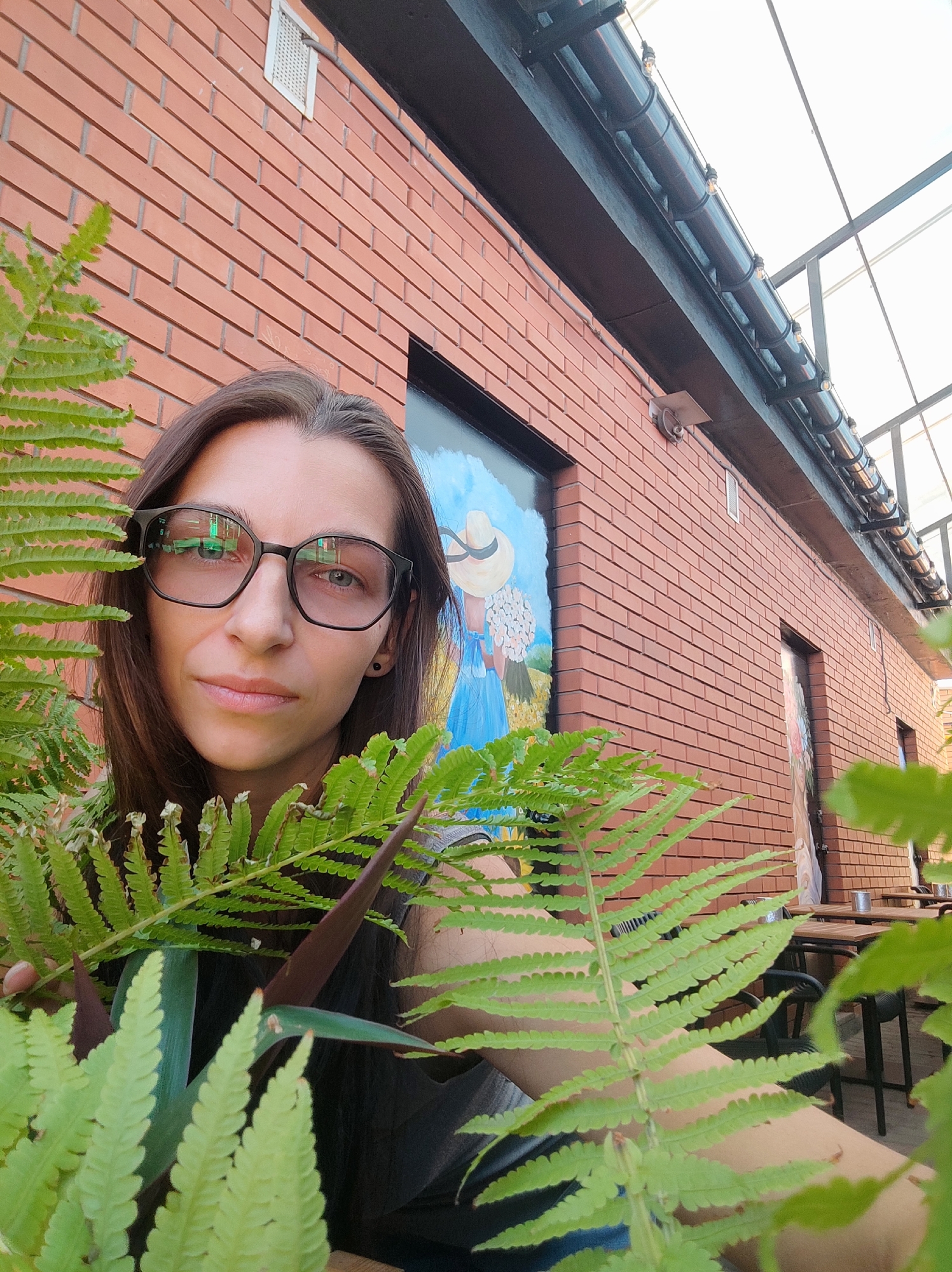An outdoor portrait is captured at an outdoor cafe. The subject, a woman wearing glasses, is partially obscured by lush green ferns in the foreground. Behind her, a brick building is visible, adorned with a mural depicting a person in a straw hat in a field. The background also shows tables and chairs, suggesting the cafe setting. The scene is lit by natural light, with a bright sky visible overhead. The location is an outdoor cafe in Kryvyi Rih, Ukraine.