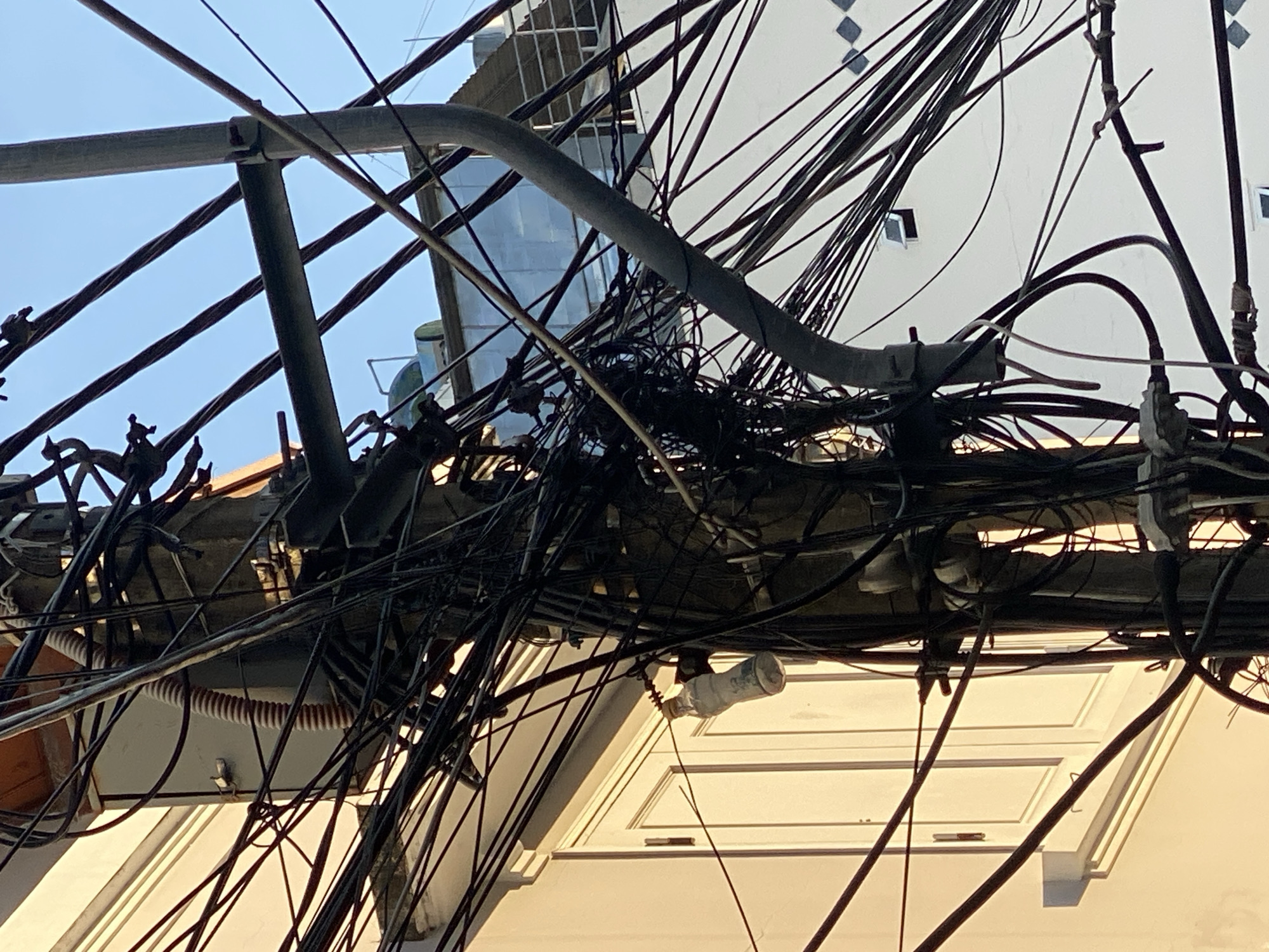 The image depicts a close-up, low-angle view of a utility pole heavily laden with tangled electrical wires and cables. The pole itself is a thick, grey cylindrical structure, appearing weathered and worn. Attached to the pole are various metal brackets and fixtures that support the dense network of black wires. Some wires are thick and appear to be power lines, while others are thinner and likely telecommunication cables.
In the background, a building with a light-colored facade and white window frames is visible. The building has multiple stories, and some architectural details, such as cornices and what appears to be an air conditioning unit or vent, can be seen. Further in the distance, another building with a white exterior and a pattern of square shapes is partially obscured by the wires. The sky above is a clear, bright blue.
A notable object hanging from one of the wires is a clear plastic bottle with a yellow cap. The bottle is partially filled with a light-colored liquid and appears to be suspended by a wire wrapped around its neck. This detail, along with the sheer density and disorganization of the wiring, suggests a somewhat haphazard or unregulated approach to utility infrastructure. The lighting indicates it is daytime, possibly late afternoon given the warm glow on the building.