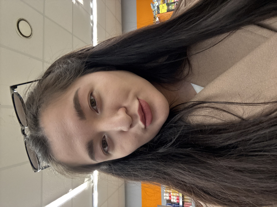 A close-up, front-facing photograph captures an adult female in an indoor retail setting within the city of Kurovskoye, Russia. The subject has long, dark brown hair with black-framed sunglasses positioned on top of her head. Her facial features include dark eyebrows, dark eyes, and lips colored with a reddish-brown matte lipstick, slightly pursed. She is wearing a light brown, possibly camel-colored, coat over a white undergarment. The background features a white tiled ceiling with visible fluorescent strip lights and at least one circular recessed light fixture. To the left, shelves stocked with various bottled or canned products are visible. To the right, an orange display or wall with a yellow price tag showing "79" is discernible.