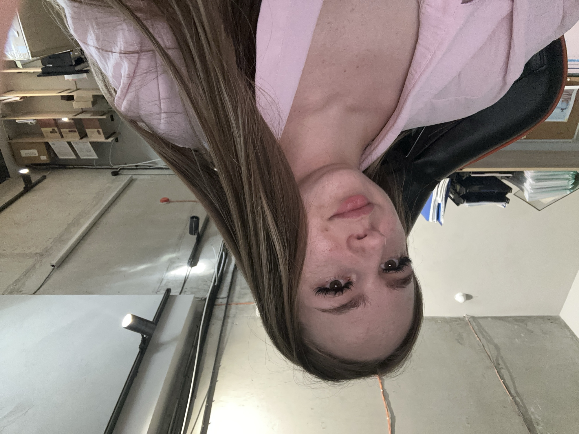 This is a low-angle, upside-down portrait of a young woman in an indoor setting. The woman has long, brown hair with lighter streaks and is wearing a light pink garment. She has noticeable eyelashes and appears to be looking directly at the camera with a neutral expression. The background suggests an unfinished or industrial space with concrete walls and exposed pipes. Shelves with boxes and what appear to be documents or books are visible in the upper left portion of the image. Lighting fixtures are also present, with one spotlight illuminating a portion of the wall. The overall impression is informal, possibly a casual selfie taken in a workshop, storage area, or similar environment. There is no visible text in the image.