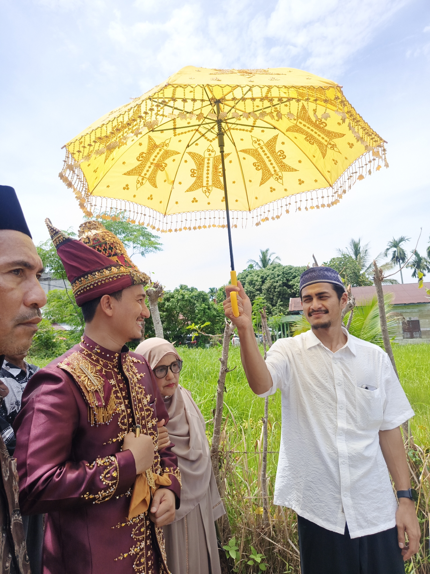In Meuliek, Indonesia, a man dressed in a traditional maroon and gold embroidered outfit and a tall, ornate headdress stands to the left, smiling and looking to his right. He is holding a gold-colored fabric in his left hand. To his right, a man in a white short-sleeved shirt and a blue peci hat holds a large, decorative yellow umbrella with intricate gold patterns and fringing. The umbrella is tilted slightly and positioned above the two men. Behind them, a woman in a light brown hijab and glasses looks forward. The background consists of lush green grass, trees, and a building with a red roof, under a bright, cloudy sky.
