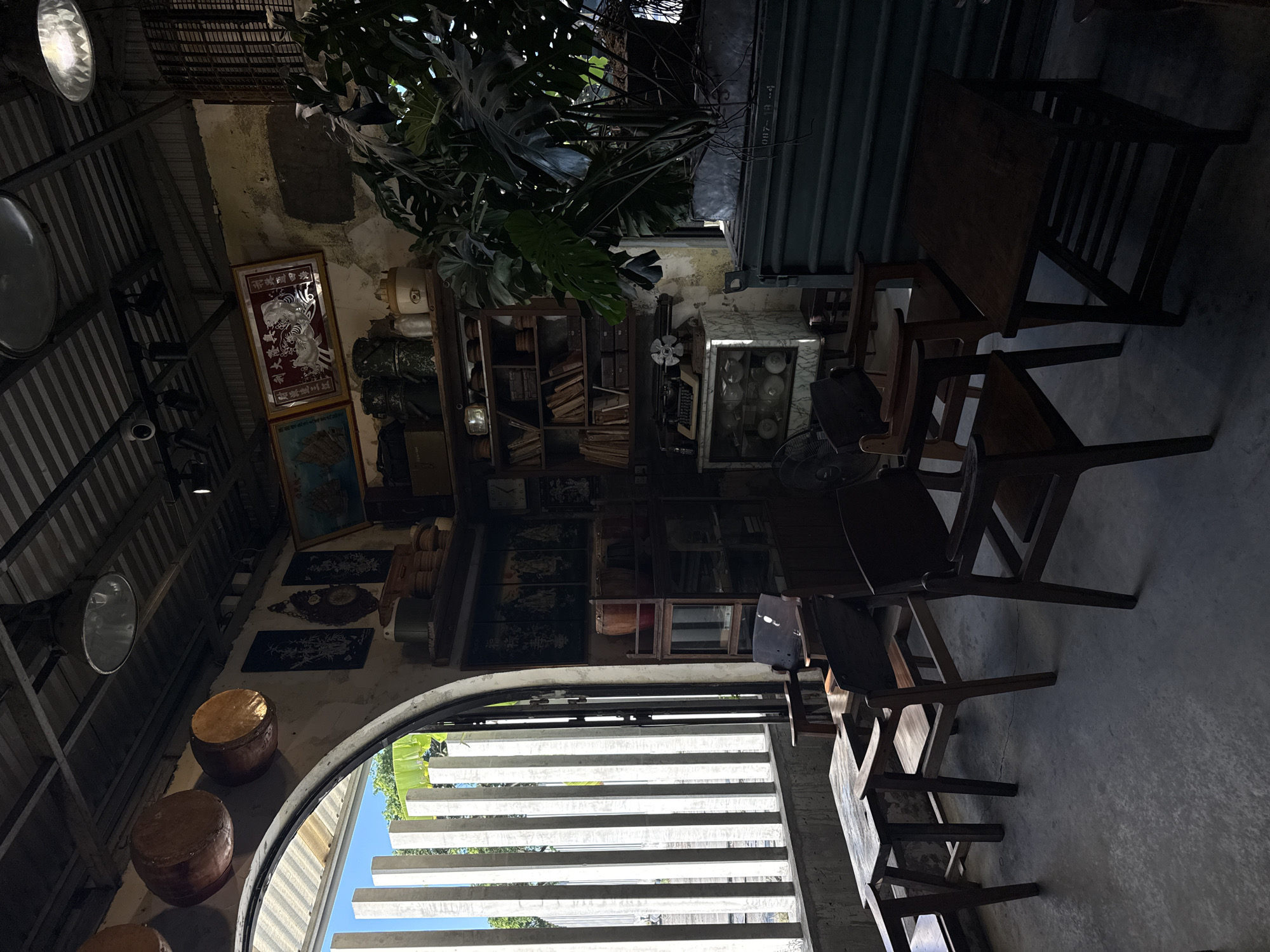 This image depicts the interior of a cafe, likely in Đà Nẵng, Vietnam, with a rustic and vintage ambiance. The cafe features a corrugated metal ceiling with industrial-style pendant lights. The walls are textured and show signs of age, with some sections appearing to be exposed brick or rough plaster.
The space is furnished with several wooden chairs and tables, arranged in a casual seating area. The furniture appears to be older, possibly antique, contributing to the vintage feel. Sunlight streams in through a large arched window with vertical slatted blinds, casting shadows across the floor and furniture.
Various decorative and functional objects are displayed around the cafe. On the wall, there are framed artworks and decorative plates. Shelves are stocked with books, containers, and other assorted items. A vintage typewriter sits on a cabinet, and a large potted plant adds a touch of greenery to the scene. In the background, a large metal structure, possibly a cabinet or storage unit, is visible. The overall impression is of a cozy and characterful cafe with a strong emphasis on a nostalgic aesthetic.