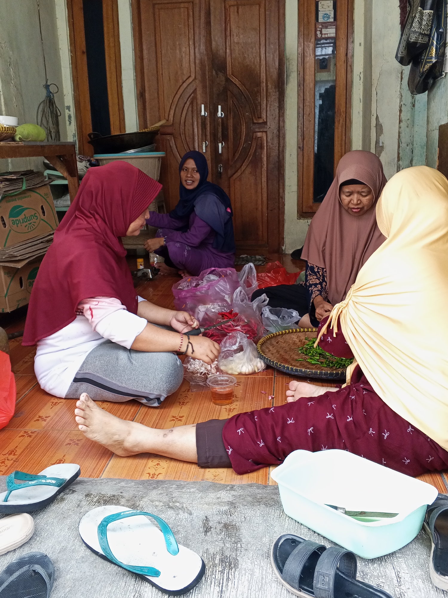 In the city of Ciruas, Indonesia, four individuals are gathered on a tiled floor in what appears to be an outdoor or semi-outdoor setting, likely a porch or common area. The scene is centered around food preparation.
On the left, a person wearing a maroon hijab and a white shirt with pink sleeves is seated, facing towards the center. They are actively engaged in chopping or preparing ingredients, with a small cup of liquid and a plastic bag of red chilies nearby. Their legs are crossed, and their feet are visible on the floor.
To their right, another person in a dark blue hijab and a purple garment is also seated, looking towards the center and smiling. They appear to be holding something in their hands, possibly also involved in food preparation.
In the middle ground, a person in a taupe hijab is seated and looking down at a woven basket filled with small green chilies. They are holding a utensil, presumably for preparing the chilies.
To the far right, a person wearing a yellow hijab is partially visible, also appearing to be involved in the food preparation. Their maroon patterned pants are visible.
The environment features a large wooden double door in the background, with details of its carvings and handles. To the left of the door, there are shelves with boxes, one of which has the "Sunpride" logo. The floor is tiled with a pattern of brown and lighter squares. Various objects are scattered on the floor, including multiple pairs of flip-flops, plastic bags, a woven basket, and a light blue plastic container. The overall atmosphere suggests a communal activity of cooking or preparing food.