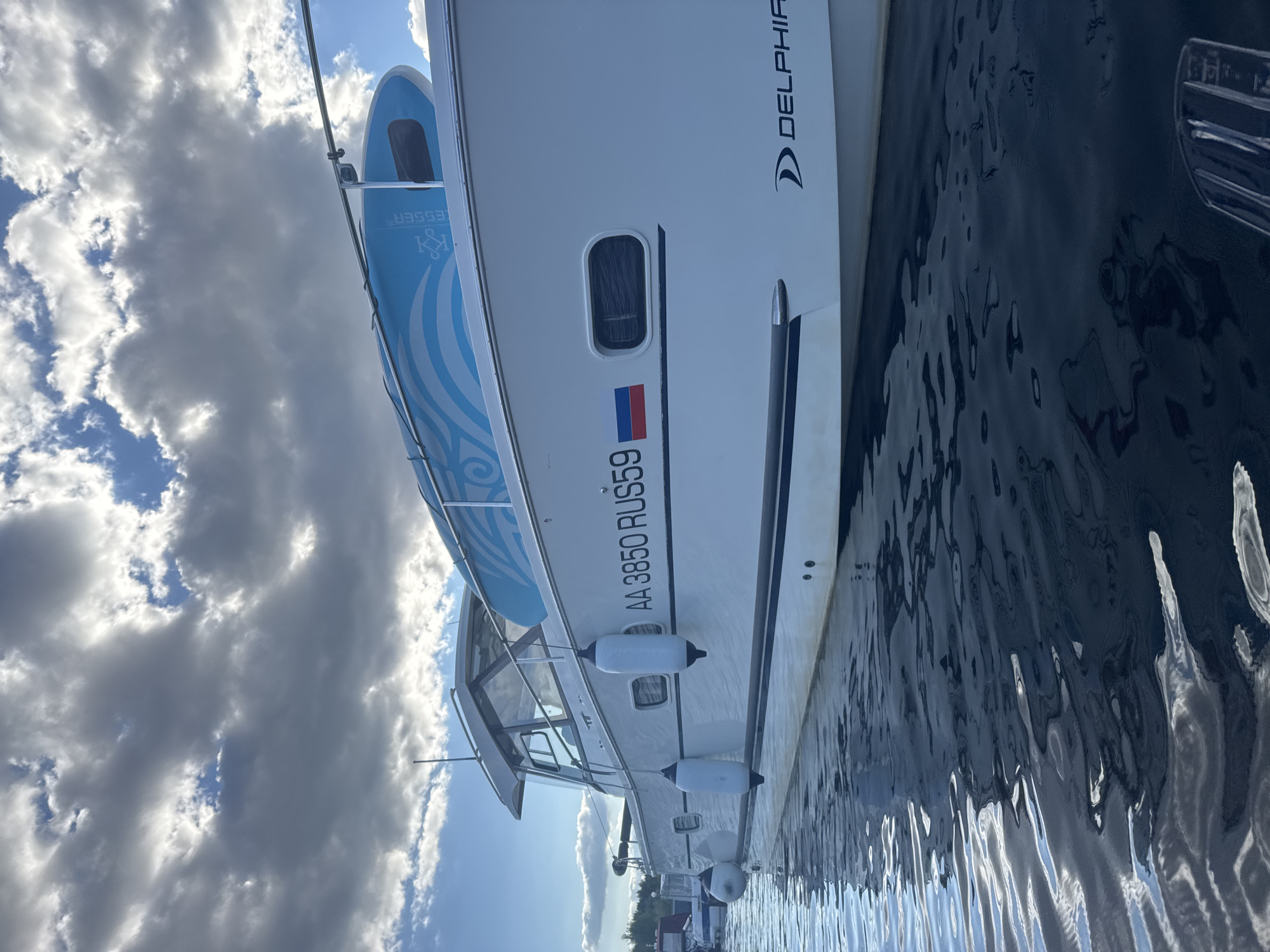 The image shows a white motor yacht named "DELPHIA" docked on the water. The yacht has the registration number "AA 3850 RUS59" displayed on its side, along with a Russian flag. A blue stand-up paddleboard with a wave-like design is secured to the roof of the yacht. The boat is positioned at a low angle, with the water reflecting the sky and clouds. The environment suggests a marina or harbor, likely in Chaikovsky, Russia, given the boat's registration. The sky is partly cloudy, with patches of blue visible between the dark clouds.