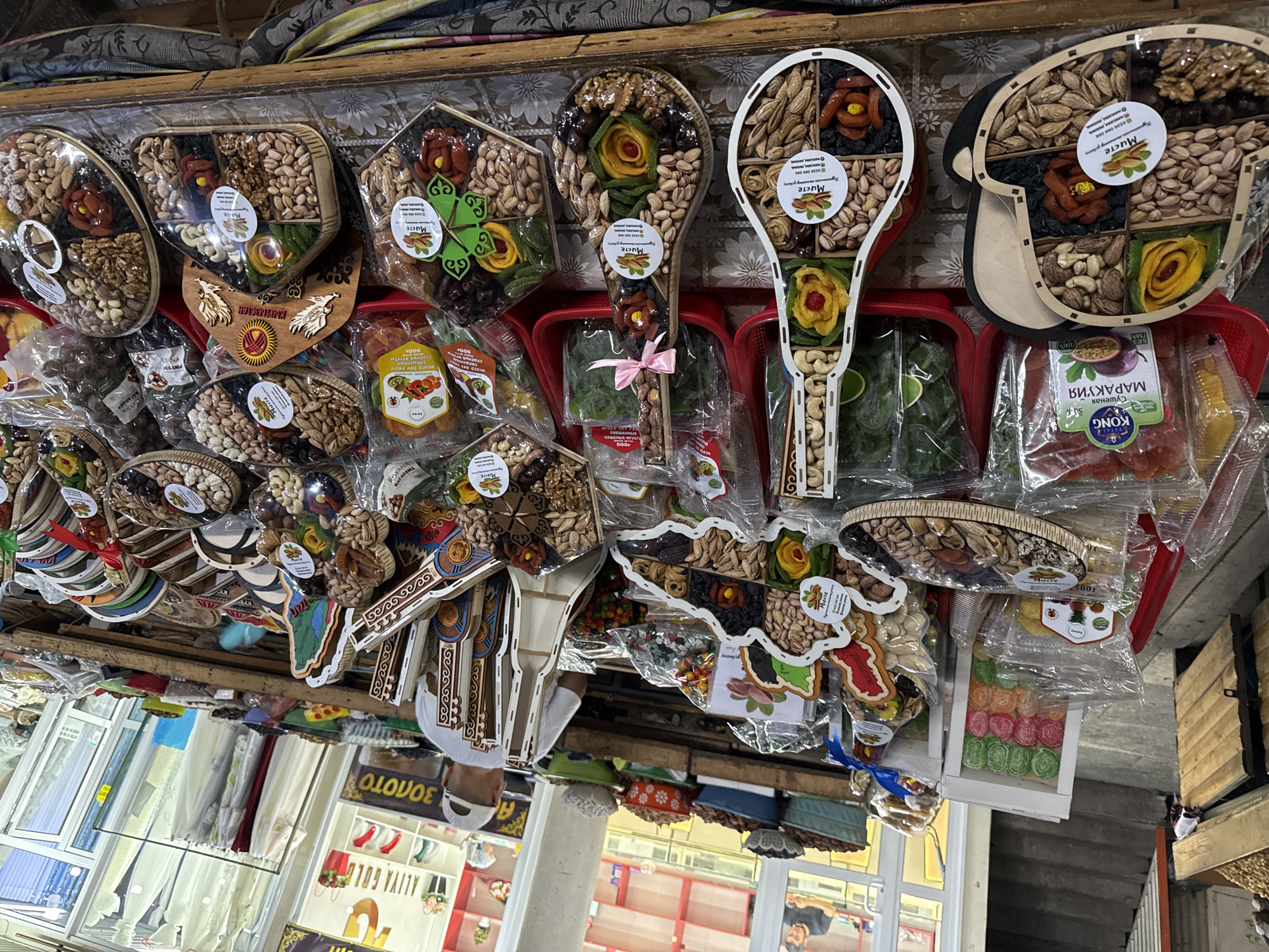 The image depicts a vibrant market stall in Manas, Kyrgyzstan, overflowing with an assortment of dried fruits and nuts. The foreground is dominated by intricately arranged gift boxes filled with these edible items. Several boxes are shaped like musical instruments, such as a guitar and a lute, showcasing a creative presentation of the products. Other boxes are heart-shaped or diamond-shaped, also filled with a variety of nuts like pistachios, almonds, and walnuts, alongside dried fruits such as apricots and dates, and what appear to be candied fruit slices. Many of these gift boxes are wrapped in clear cellophane, revealing the colorful contents within.
In the background, the market environment is visible. Wooden crates and shelves hold larger quantities of nuts and dried fruits. A man wearing a white baseball cap and a white t-shirt is seen from behind, attending to the stall, suggesting a commercial setting. The market appears to be indoors or under a covered structure, with stalls and displays extending into the distance. The lighting suggests it is daytime, with natural light filtering in from openings. Signs and labels in Cyrillic script are visible on some of the packaging and in the background, indicating the local language and context of the market. The overall impression is one of a bustling marketplace offering a wide selection of traditional and attractively packaged dried goods.