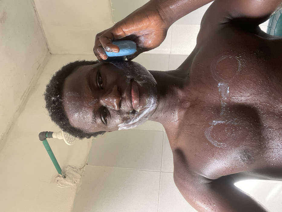 A young adult male, shirtless and wet, is shown taking a selfie in a bathroom located in the city of Mosafejaw, Nigeria. He has dark, short, curly hair and is smiling directly at the camera. White lather is present on his face, specifically on his cheeks and chin, and two distinct circular patterns of lather are visible on his wet chest. His left hand holds a blue bar of soap to his left ear. In the background, a white showerhead is mounted on a light-colored wall via a green pipe, with the lower wall section tiled in a lighter shade. The ceiling above shows signs of discoloration.