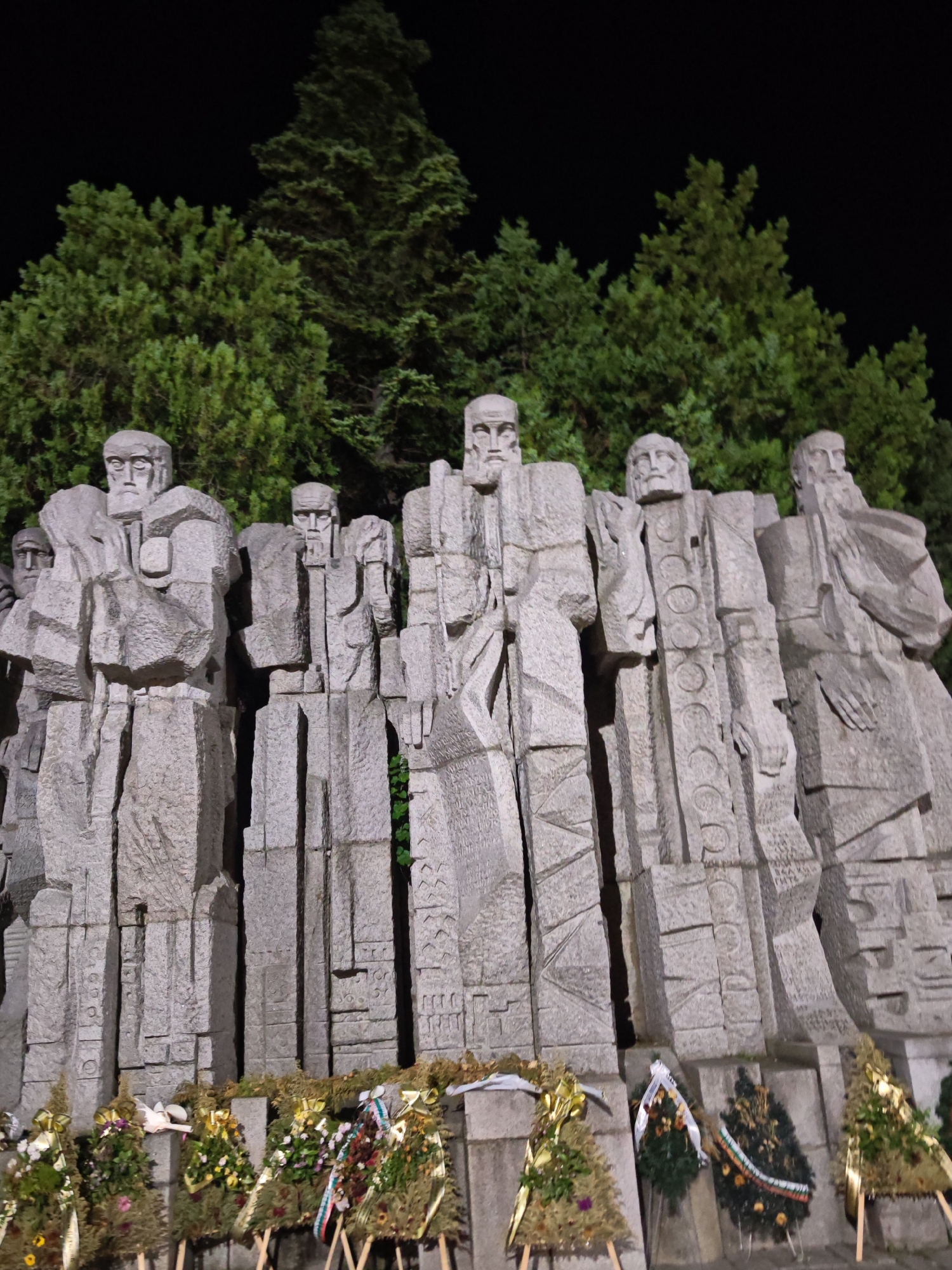 The image captures a nighttime view of the "Monument to the Golden Age" (also known as the "National Monument 'Golden Age'") in Veliki Preslav, Bulgaria. The central focus is a large, monumental stone sculpture comprising at least seven stylized human figures carved from light-grey stone. These figures are presented in a blocky, angular, and somewhat cubist style, depicting bearded male individuals in robes with abstract facial features. Notable details include a figure on the far right with a long, flowing beard, and a central figure adorned with a vertical series of circular medallions carved into his attire.
At the base of the monument, several commemorative wreaths are arranged on individual wooden stands. These wreaths are composed of evergreen boughs, various flowers, and decorative ribbons, with some ribbons displaying the colors white, green, and red, indicative of the Bulgarian national flag. The background features dense evergreen trees silhouetted against a completely dark, black night sky. The scene is illuminated by artificial lighting, which highlights the texture and form of the stone monument and the greenery, creating a stark contrast with the unlit sky. No human presence or activity is visible within the frame.