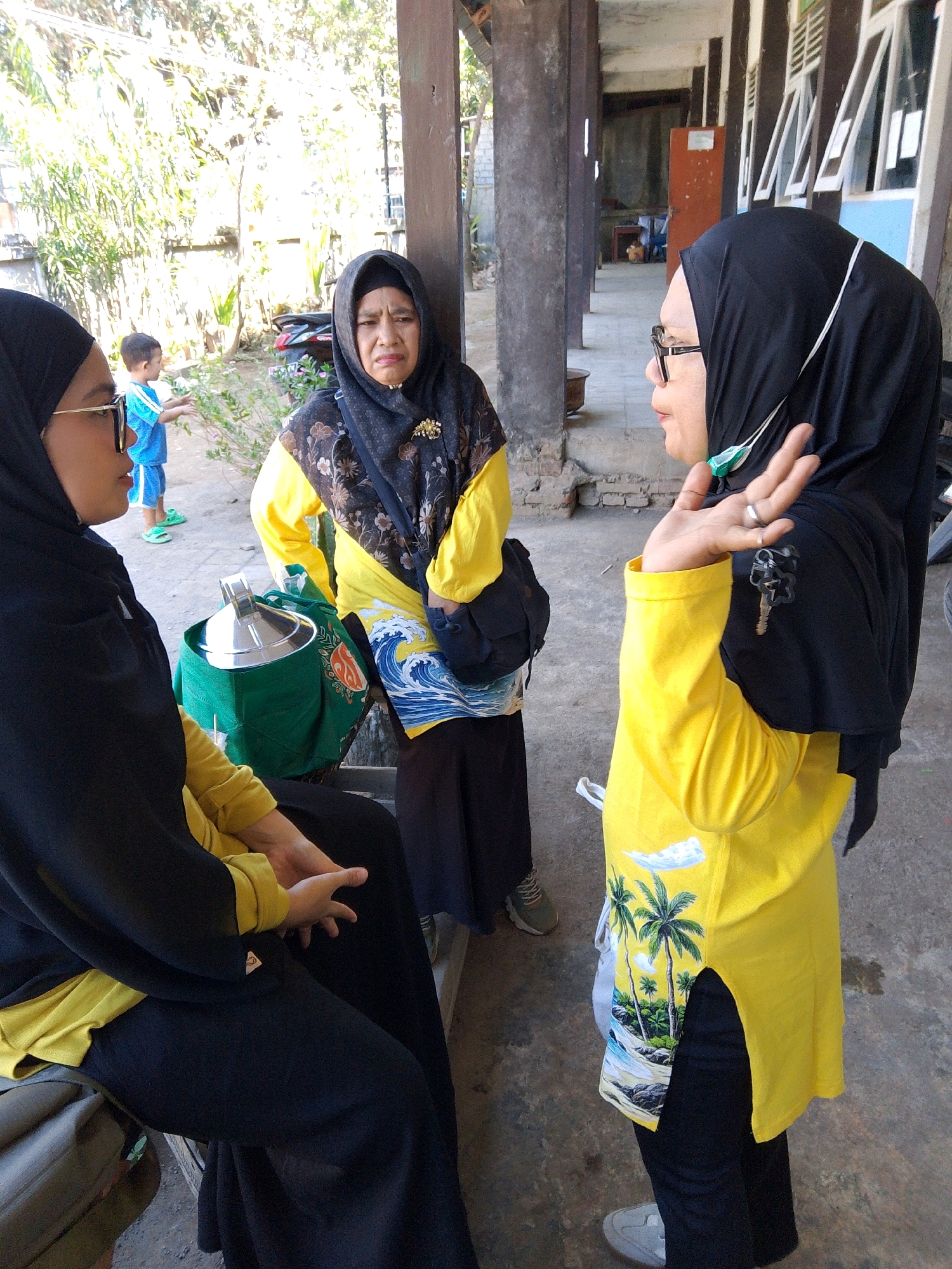 In Kuripan Utara, Indonesia, three women and a young boy are seen in an outdoor, possibly school or community setting. One woman, wearing a yellow shirt and black hijab, is speaking with another woman in a black hijab, who is gesturing with her hand and wearing glasses. The second woman is also wearing a black hijab and glasses. The third woman, wearing a black hijab and a patterned top with yellow sleeves, appears to be looking down with a displeased expression. A young boy in a blue shirt and shorts stands in the background, facing away from the camera. They are situated on a paved surface with some greenery visible. A large green bag with a silver container is placed on the ground next to one of the women. The environment suggests a sunny day, with shadows cast by the overhead structure and surrounding foliage.
