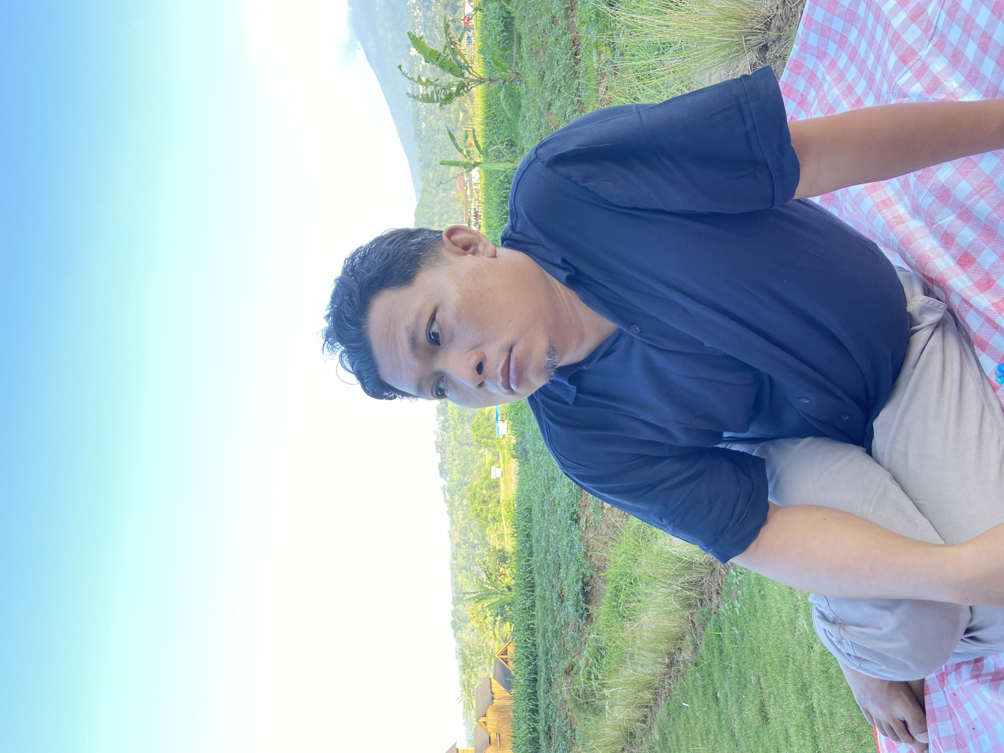An outdoor, medium shot, portrait-style photograph shows a man sitting on a red and white checkered blanket in a grassy field. The man is facing to his left, looking into the distance with a neutral expression. He has short, dark hair and is wearing a dark blue polo shirt and light-colored pants. The background features a lush green landscape with rows of crops, some small wooden buildings, and rolling hills under a clear blue sky with a hint of clouds near the horizon. The location is identified as Kesiman, Indonesia.