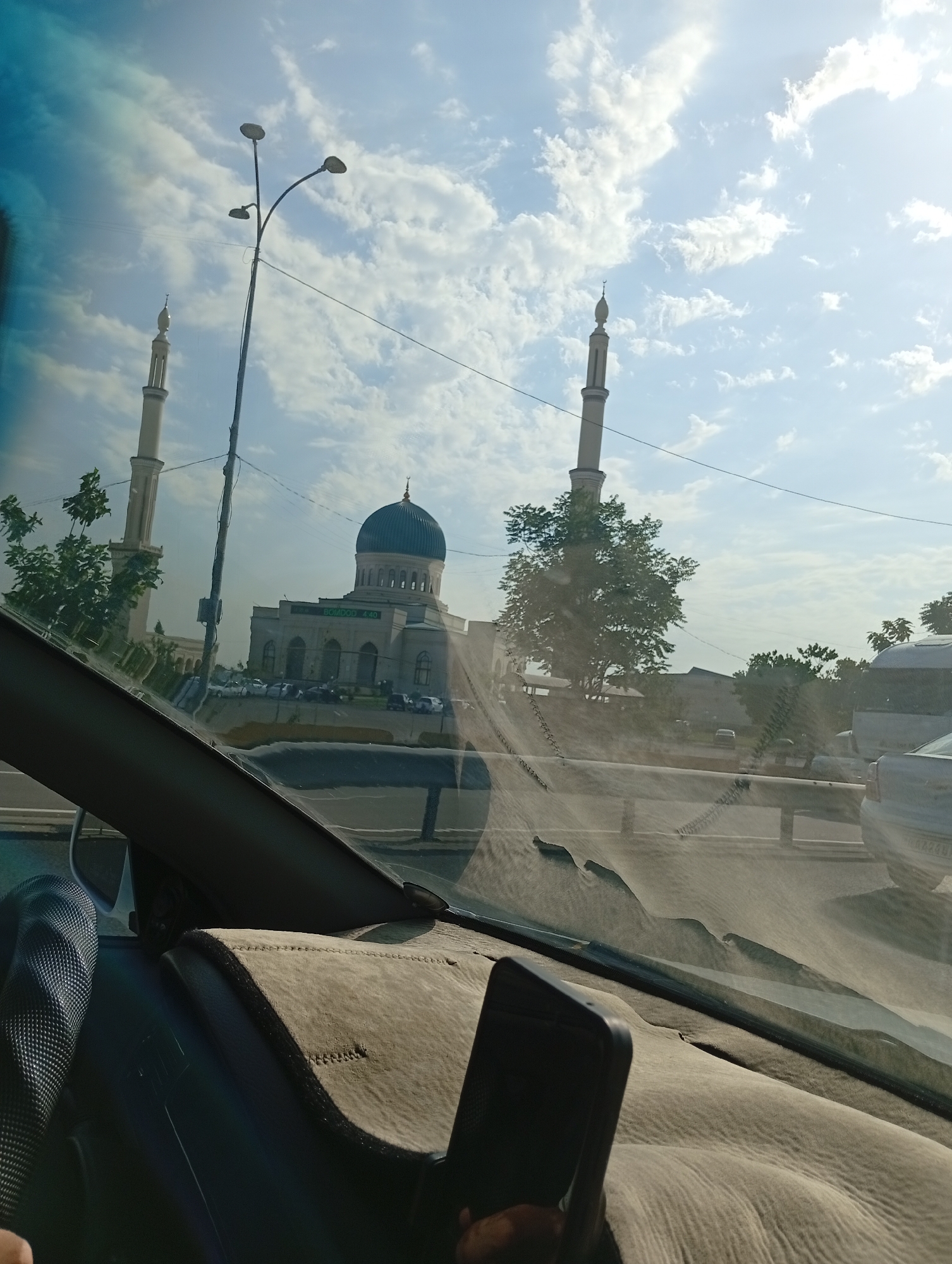 The image captures a view from inside a vehicle, looking through the windshield towards a mosque in Ohangaron, Uzbekistan. The mosque, with its large dome and two minarets, is the central focus of the scene. The sky is partly cloudy with bright sunlight. Trees line the path leading to the mosque, and several cars are parked in front of it. A street lamp with three lights stands to the left of the mosque. The interior of the car, including the dashboard, steering wheel, and a mobile phone, are visible in the foreground. The overall impression is a sunny day in a city, with the mosque serving as a prominent landmark.