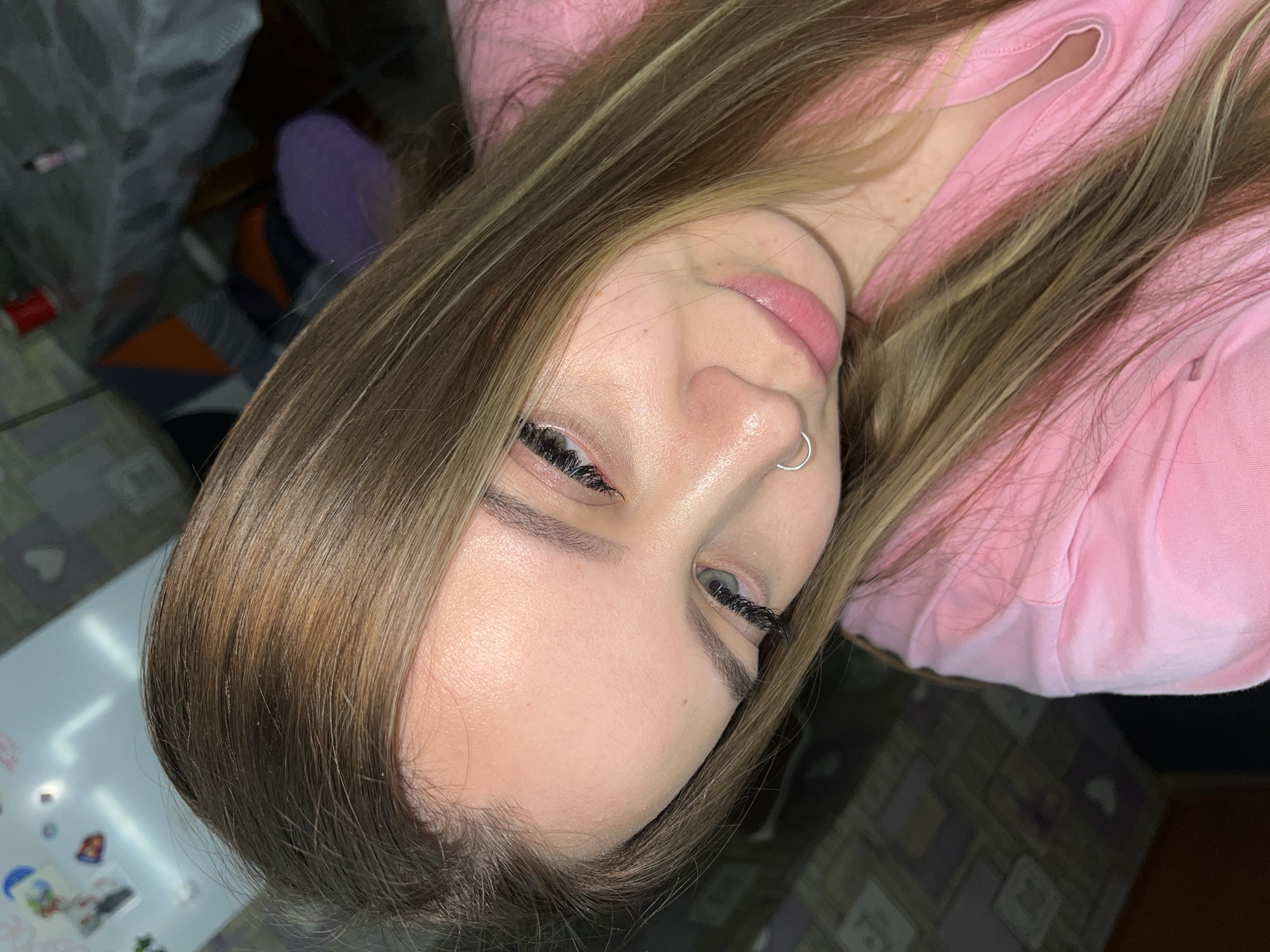 A close-up, slightly rotated image shows a light-skinned person with light brown hair and a silver nose ring. The person is wearing a pink shirt. The background includes a table surface with stickers and other objects, and a patterned surface. The image provides no information about the city of Port-Katon, Russia.