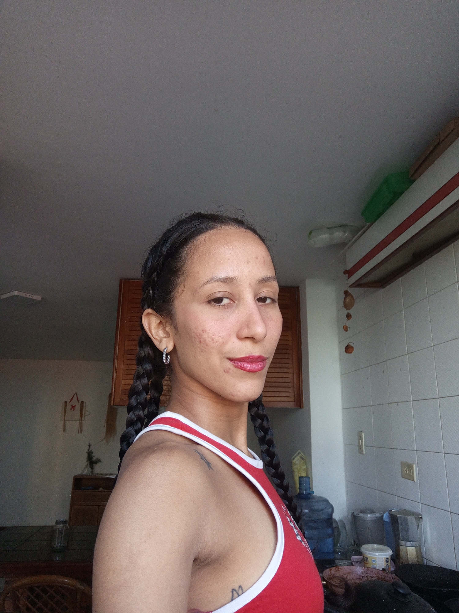 A woman with dark braided hair and light-colored hoop earrings is photographed from the chest up, looking to her right. She is wearing a red tank top with white trim. Her skin has some blemishes. The background appears to be a kitchen, with tiled walls, cabinets, and a water cooler visible. There are also some items on a shelf above the tiles, including a green object and some dried decorations. The lighting suggests it is daytime. This scene is likely taking place in Porlamar, Venezuela, given the context.
