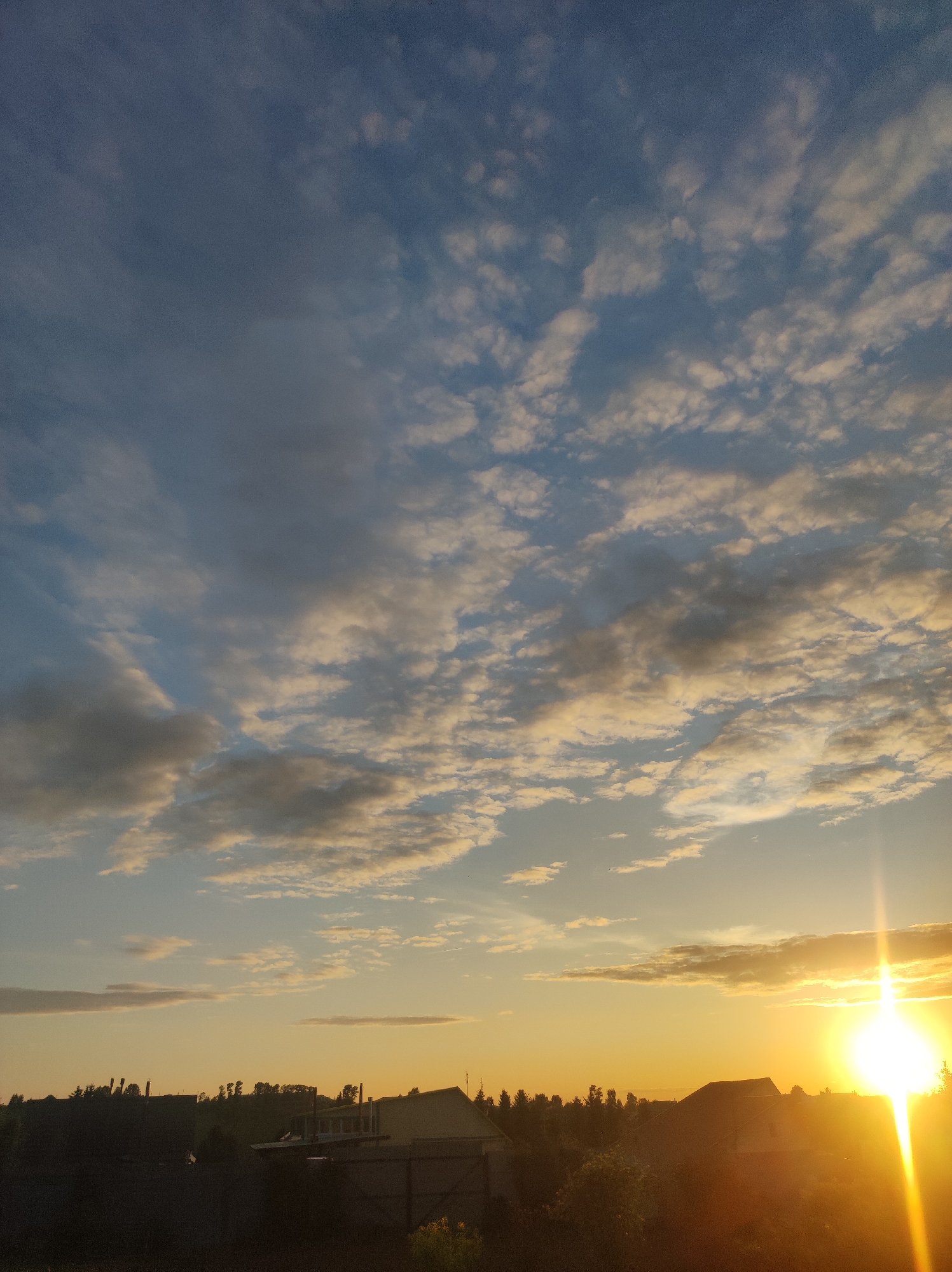 The image captures a rural or suburban landscape at either sunrise or sunset in Фоки, Russia. The sky occupies the majority of the frame, displaying a gradient from a dark blue-grey at the upper left to bright orange and yellow tones at the right horizon. Scattered altocumulus clouds, varying in shade from grey to white and illuminated gold, are present throughout the sky, becoming denser and brighter towards the sun. An intense, star-shaped sun flare with prominent rays extends from the far right of the frame, near the horizon, casting a strong yellow-white light that partially obscures the view. The lower portion of the image is dominated by a dark silhouette of residential structures, primarily houses with visible roofs, and dense tree lines and vegetation. No individuals or specific activities are discernibly present within the scene.