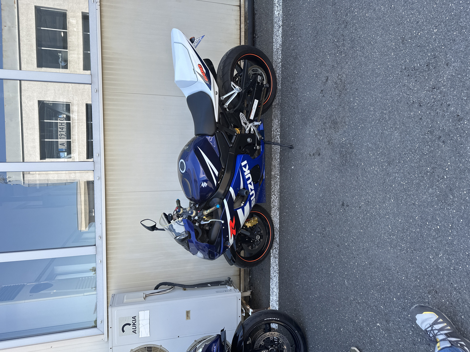 The image shows a parked Suzuki motorcycle, predominantly blue with white and red accents, with its kickstand down. The motorcycle is positioned on an asphalt surface next to a light-colored, textured wall. An air conditioning unit is visible to the left of the motorcycle, with an "AUKIA" logo on it. In the background, a building with reflective windows is visible, and a license plate with the characters "A 1914 HO" is reflected in one of the windows. Part of another motorcycle, a person's leg and foot wearing a sneaker, and the pavement are also present in the lower portion of the image. The scene is illuminated by natural daylight, casting shadows consistent with an outdoor environment.
