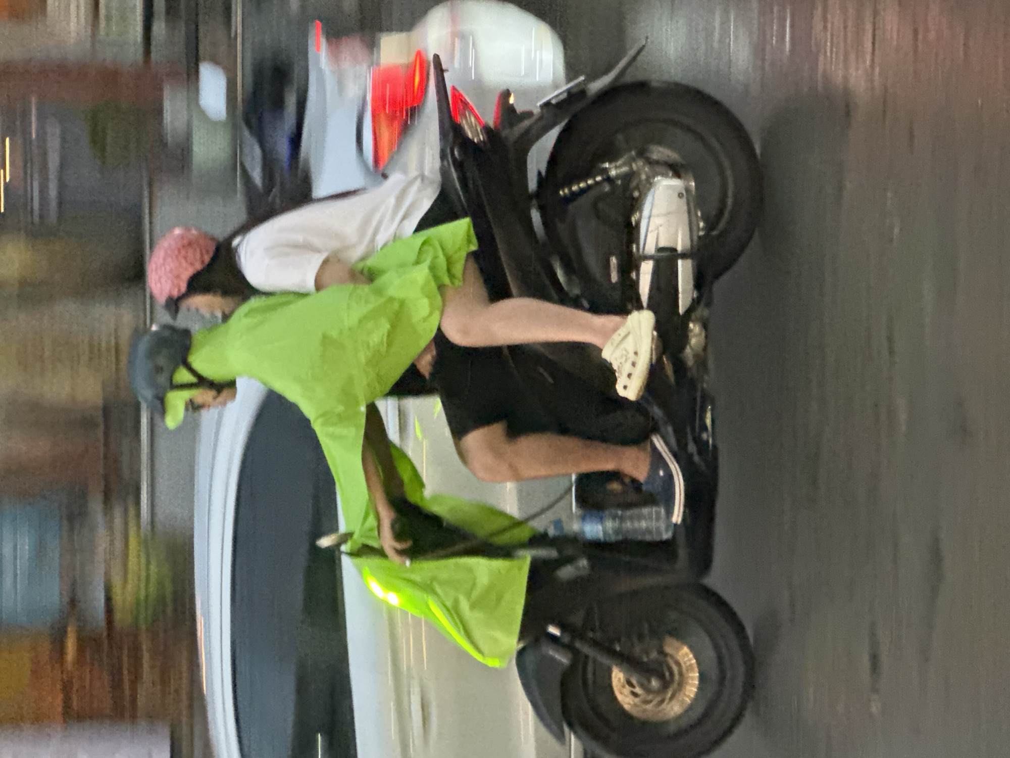 In the city of Bắc Nha Trang Ward, Vietnam, two individuals are riding a black motorcycle. The driver wears a grey helmet and a bright green raincoat. The passenger wears a white shirt and a pink knit cap. The motorcycle has a white car passing it. A bottle of water is on the floorboard of the motorcycle. The road appears wet.