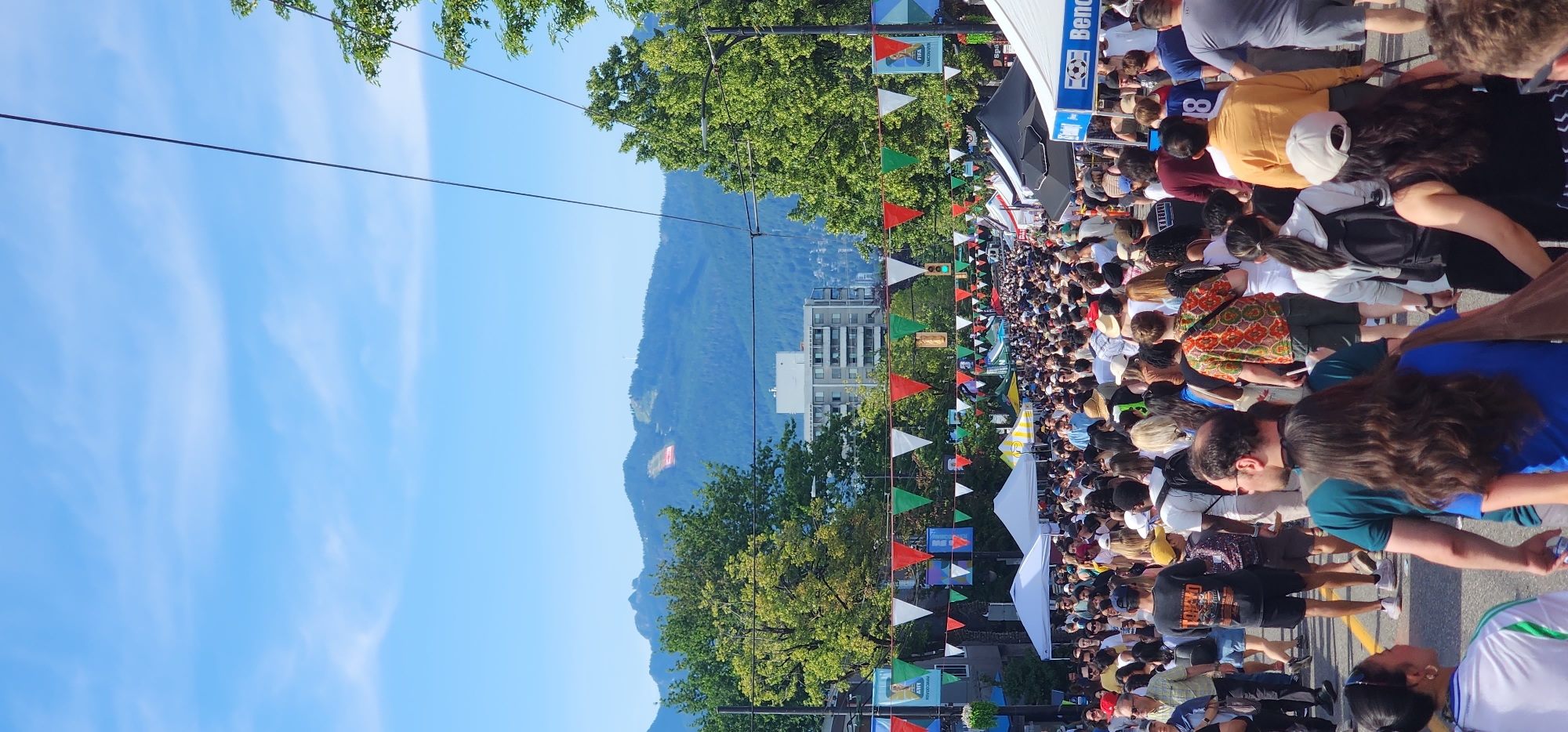 A bustling street festival is captured from a low-angle perspective looking down a crowded street lined with tents and vendor stalls. A dense throng of diverse people walks along the street, many dressed in casual summer attire. Colorful triangular flags and banners, some bearing logos like "FIFA VANCOUVER," are strung across the street overhead. Beyond the festival, a cityscape with modern buildings is visible, backed by lush green trees and a mountain range under a bright blue sky with scattered white clouds. The overall scene suggests a lively outdoor event on a sunny day.