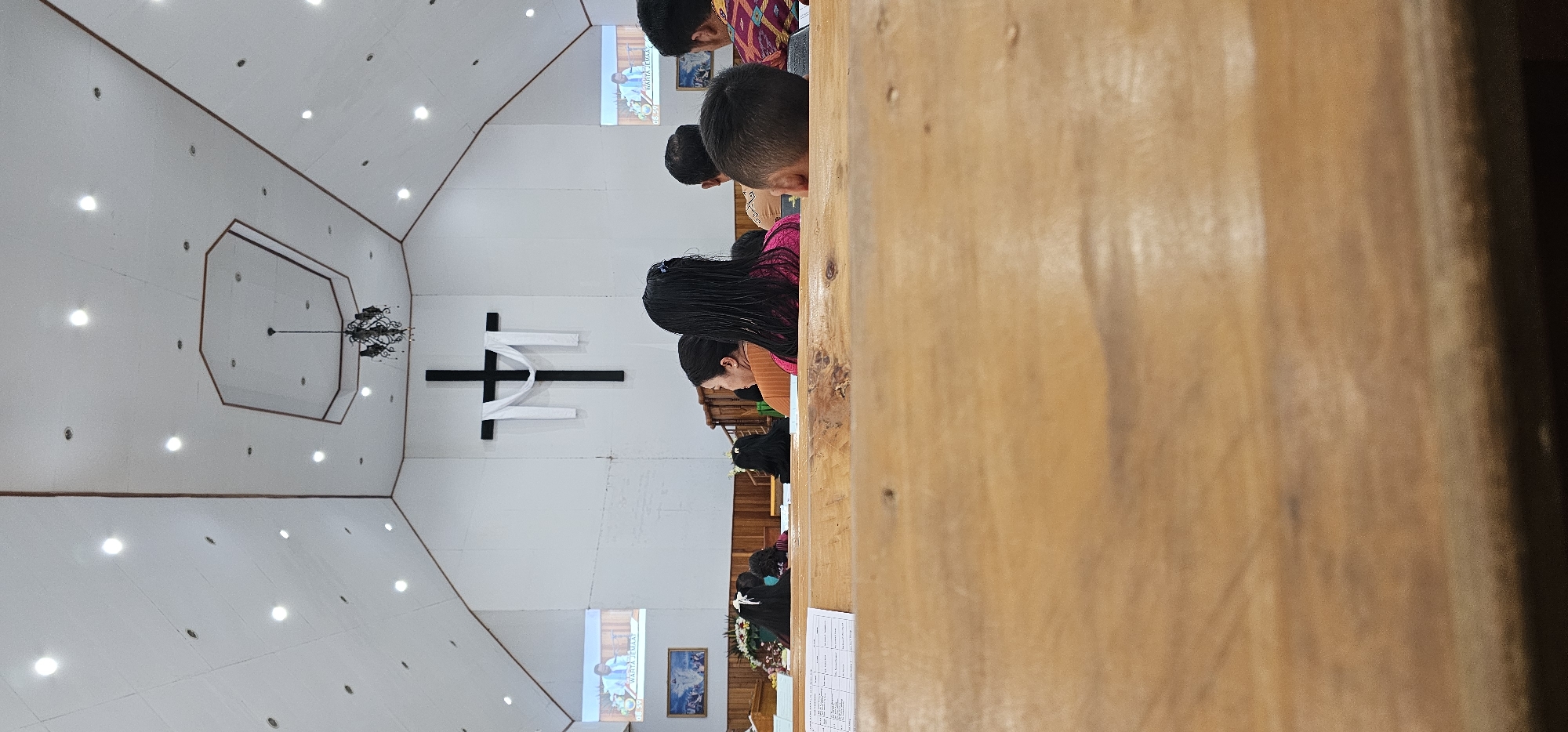 The image depicts the interior of a church, likely located in Puncak Jaya, Indonesia, as indicated by the context. The scene is viewed from a low angle, focusing on the wooden pews in the foreground, with the back of some attendees' heads visible. In the middle ground, several individuals are seated, their backs to the camera, facing a large black cross adorned with white cloth, mounted on the white wall at the front of the church. Two projection screens are visible on either side of the cross, displaying images. A chandelier hangs from the octagonal ceiling feature. The overall ambiance suggests a religious service in progress.