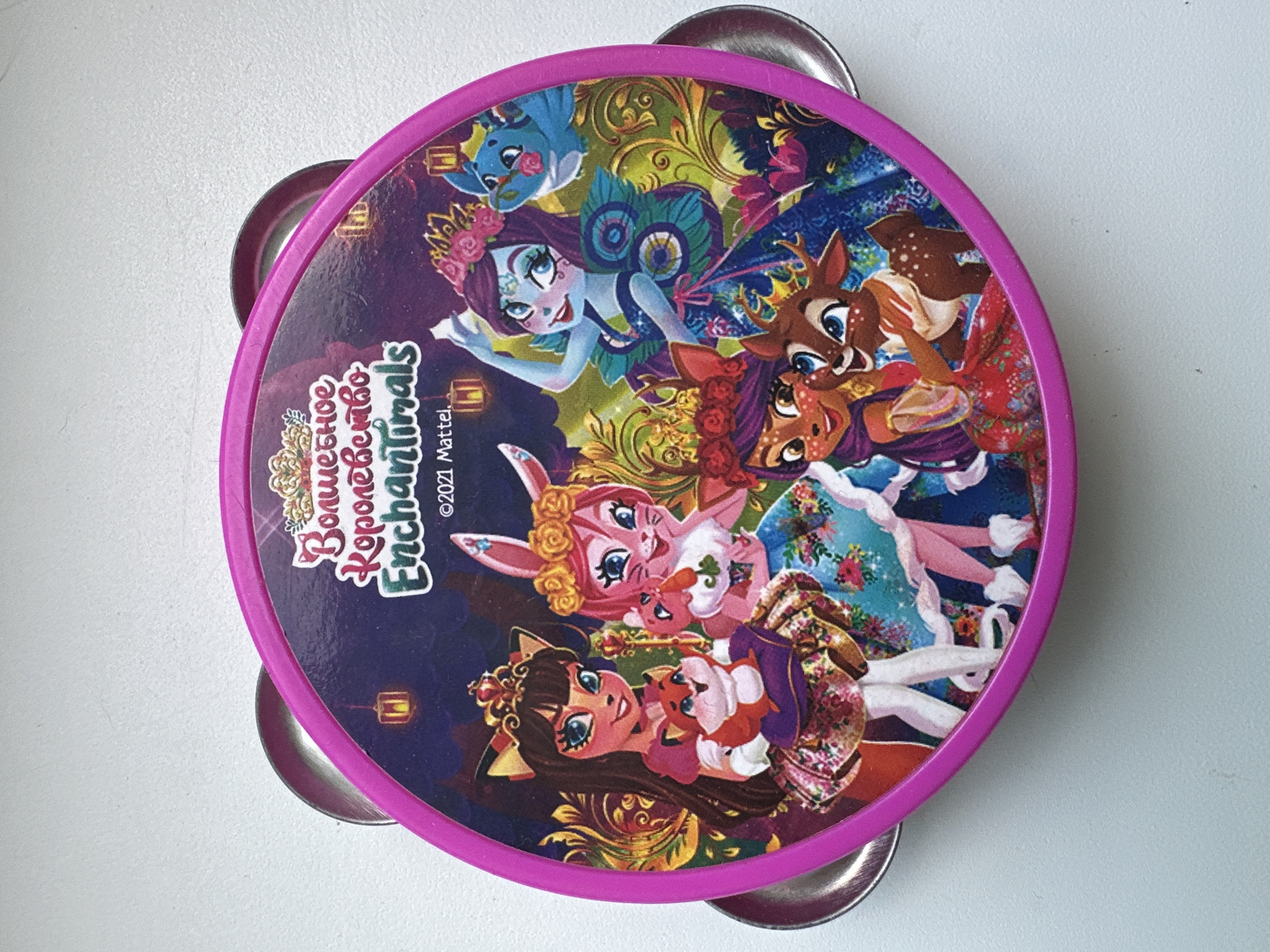 This media file shows a top-down view of a circular tambourine with a bright pink rim and silver jingles. The surface of the tambourine features a vibrant illustration with a dark purple background. The artwork depicts several anthropomorphic characters, including what appears to be a blue fairy with butterfly-like wings, a deer with large blue eyes, a rabbit with pink fur, and a girl with brown hair and cat-like features. They are all dressed in colorful, patterned clothing and surrounded by ornate floral and swirling designs. One character is holding a small red and white creature.
Visible text on the tambourine includes "Волшебное Королевство Enchantimals" in a decorative font, which translates to "Magic Kingdom Enchantimals." Below this, "©2021 Mattel" is printed, indicating the copyright and manufacturer. The scene appears to be whimsical and fantastical, with no specific time of day or weather cues indicated. The setting is a plain, light-colored surface, likely a table or floor, in what is identified as Ufa, Russia. The characters' expressions are generally cheerful and engaging, contributing to the playful and magical theme of the artwork.