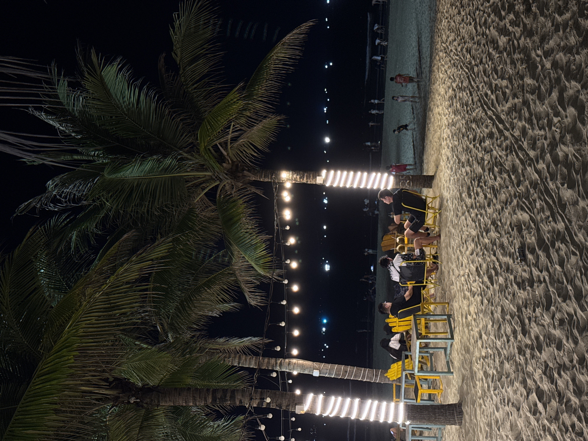 This is a nighttime photograph of a beachside setting in Đà Nẵng, Vietnam. The scene is illuminated by numerous string lights, creating a warm and inviting ambiance.
**Subjects:**
* **People:** Several individuals are visible. In the mid-ground, a group of people are seated at small, simple tables with yellow and blue chairs, appearing to socialize. One person in a black shirt is prominently seen in the foreground, seated. Further in the background, near the water's edge, more figures can be discerned walking or standing on the beach.
* **Objects:** Two prominent palm trees with full fronds dominate the left side of the frame. Their trunks are wrapped with glowing white string lights. Additional string lights are strung horizontally, creating a canopy of light. Small, square tables and simple chairs (some yellow, some blue) are set up on the sand.
* **Scene:** The setting is a sandy beach at night. The sand shows ripples and texture, indicative of footprints or natural patterns. The dark expanse of the sea or sky fills the background, punctuated by distant, scattered lights.
**Activity or event taking place:**
People are gathered at what appears to be an outdoor, casual beachfront cafe or lounge area. They are seated, likely enjoying drinks or conversation, and generally socializing in a relaxed evening environment. Other individuals are seen strolling along the beach in the distance.
**Notable details:**
* **Time of day:** Clearly nighttime, evidenced by the dark sky and reliance on artificial lighting.
* **Weather:** The conditions appear calm, suitable for an outdoor gathering, with no visible precipitation or strong wind.
* **Location cues:** The palm trees, extensive sandy area, and distant water lights strongly suggest a tropical or coastal beach setting. The specific location is Đà Nẵng, Vietnam.
* **Emotions:** The lighting and grouping of people suggest a relaxed, social, and enjoyable atmosphere.
* **Visible text:** No visible text or signage is apparent in the image.