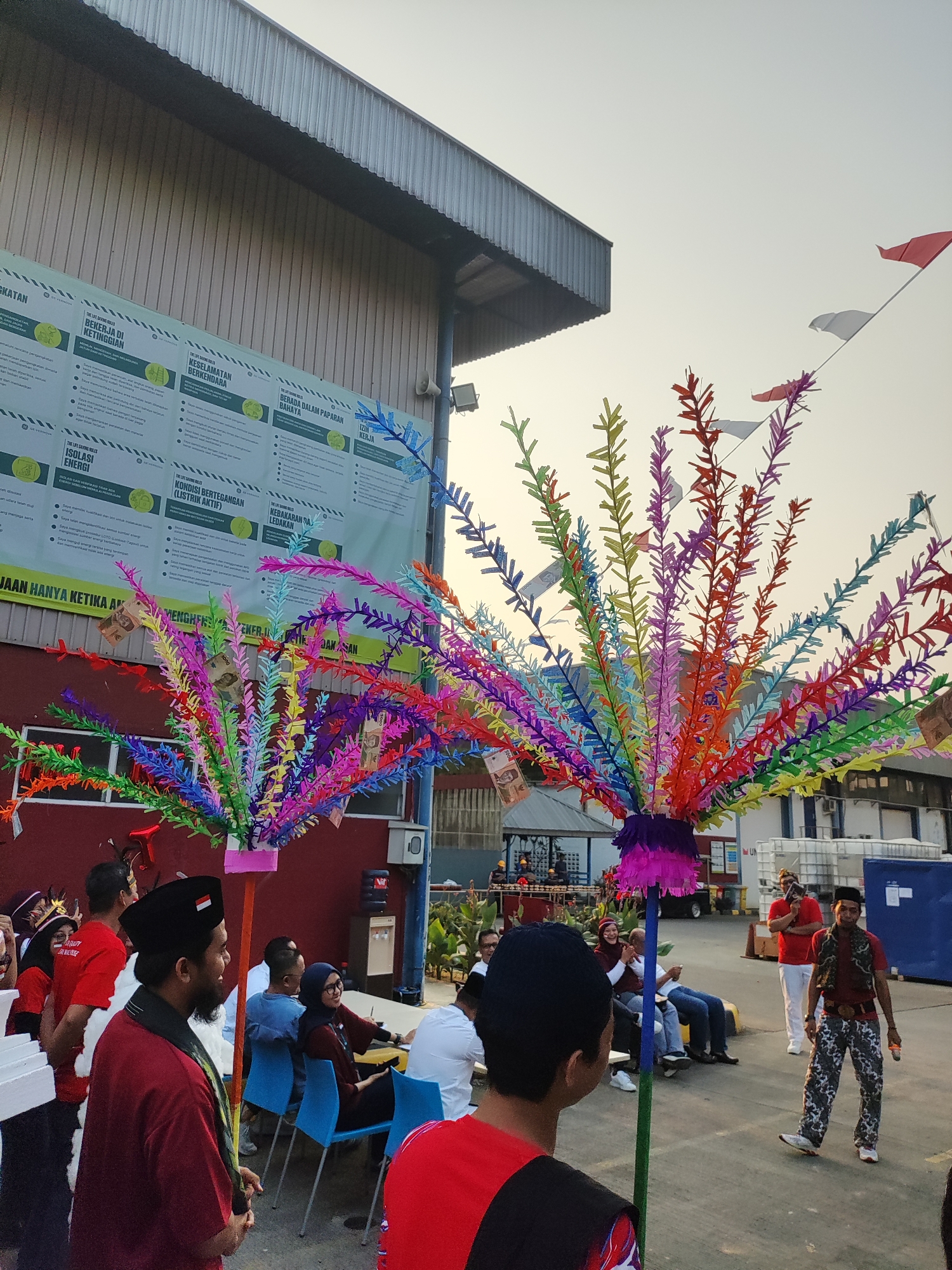 The image depicts an outdoor scene in East Jakarta, Indonesia, likely during an event or celebration. In the foreground, two people are visible, one wearing a red shirt and a black peci hat, facing to the left and holding a decorative pole with colorful, feather-like decorations adorned with Indonesian currency. Another person, from behind, is wearing a red shirt and a black peci hat.
In the middle ground, several people are seated and standing. Some are watching the proceedings, while others are engaged in conversation. One individual, in the center right, is wearing a red shirt and camouflage pants, holding a microphone and appearing to be speaking. Two colorful, decorative trees with feather-like ornaments are prominently featured in the center of the image. In the background, a large industrial building with a corrugated metal roof is visible, along with a white banner displaying text in Indonesian. Flags are strung across the top of the scene. The overall atmosphere suggests a community gathering or festival.