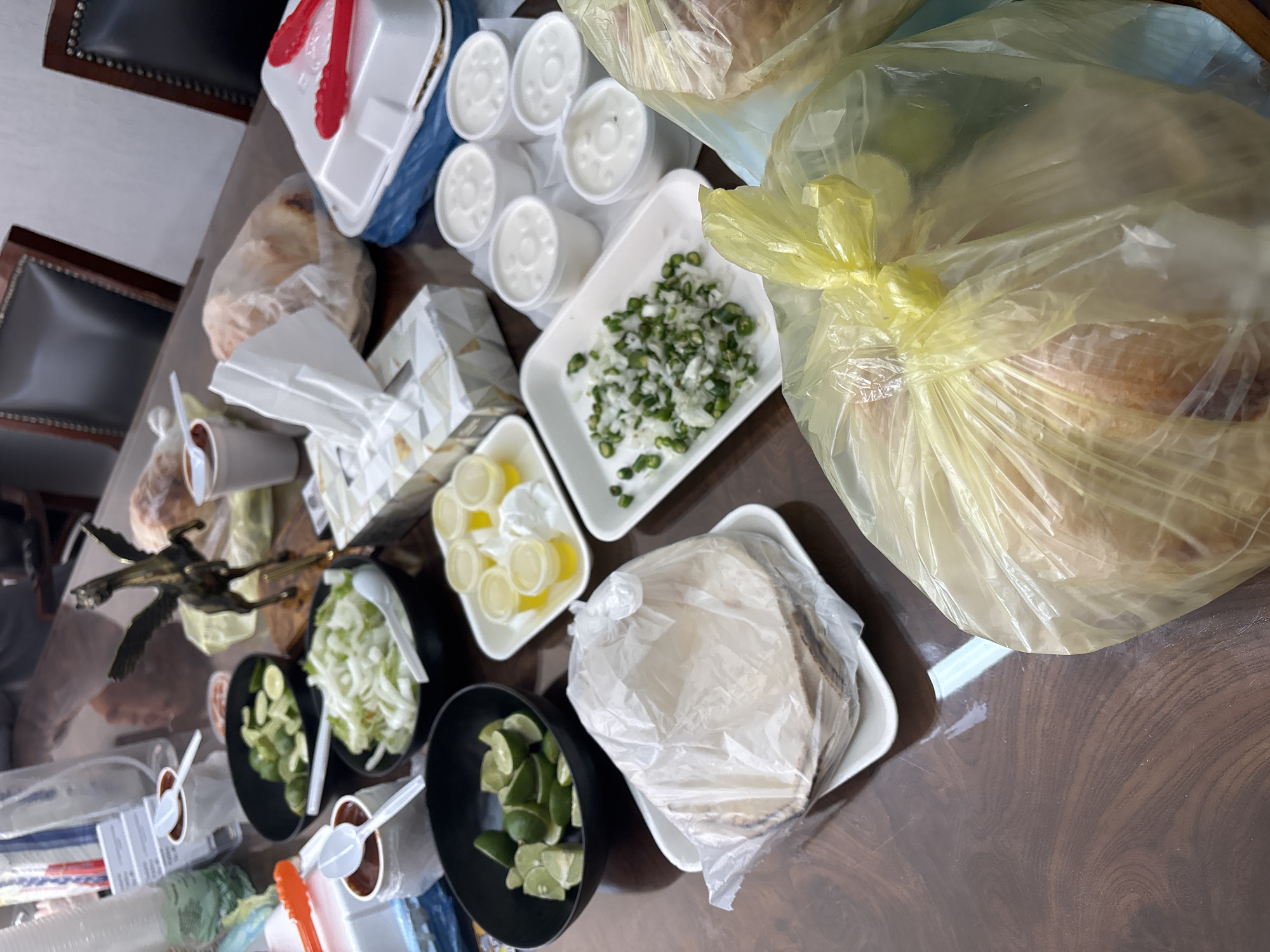 On a dark wooden table, numerous food items are arranged, suggesting a gathering or meal preparation. A large transparent plastic bag filled with bread rolls dominates the upper right. To its left is a tray of chopped onions and green chilies. Six small white cups are clustered together. There is also a white styrofoam container with red tongs placed on top of a blue plastic bag. A tissue box sits in the middle. Several black bowls contain slices of lime and shredded lettuce. Small cups of what appear to be red salsa, each with a white plastic spoon, are also on the table. A shiny metal eagle is near the limes and lettuce.
Given the type of food items such as the sliced limes, onions, tortillas, salsa, and a prevalence of traditional Mexican dishes is being suggested by the presence of such condiments and food items, this location is likely Puebla, Mexico. No people are visible.
