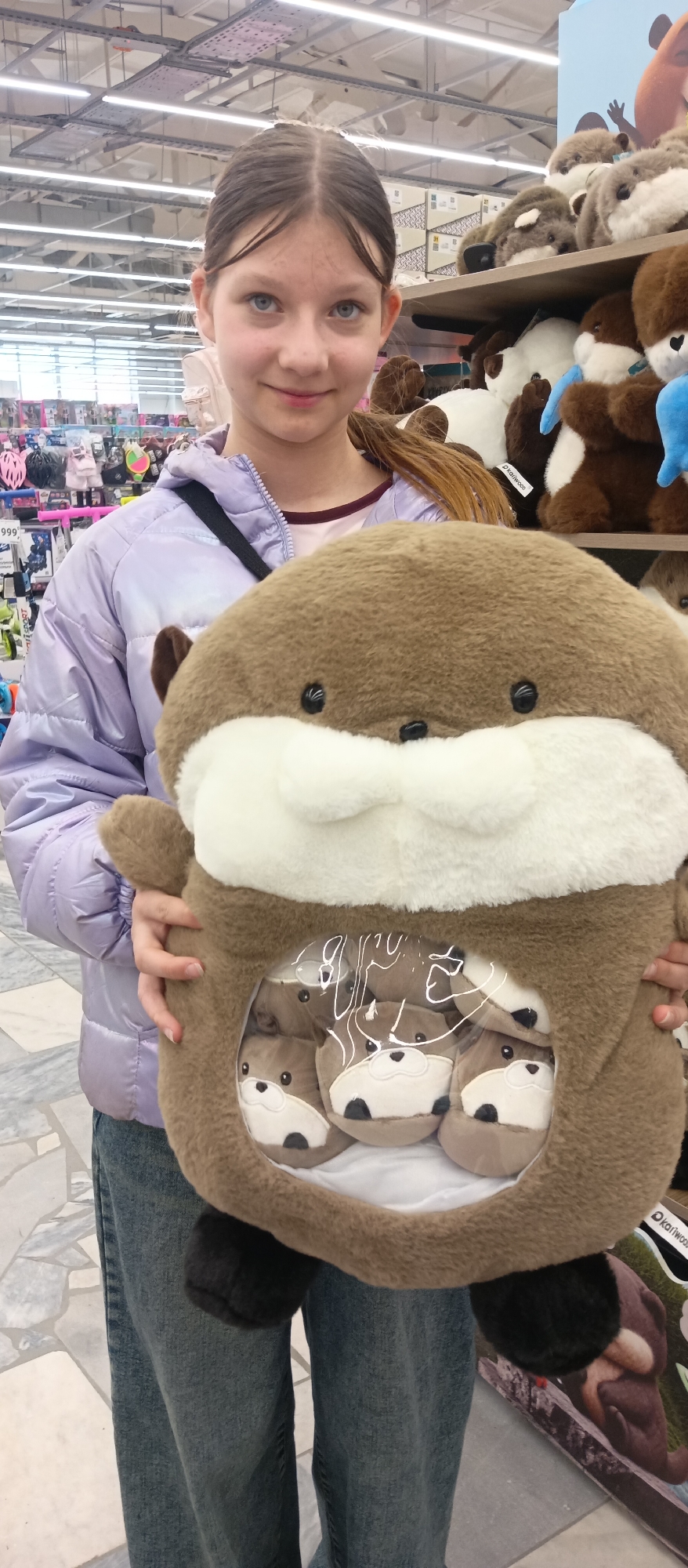 A young girl with brown hair, blue eyes, and fair skin stands in what appears to be a retail store, holding a large, round, brown plush toy. The toy has a white patch on its chest and a clear plastic window on its stomach, revealing several smaller plush toys inside. The girl is wearing a light purple, quilted jacket and jeans. In the background, shelves are stocked with a variety of merchandise, including more plush toys. Overhead, fluorescent lights illuminate the store. The location appears to be a retail store.