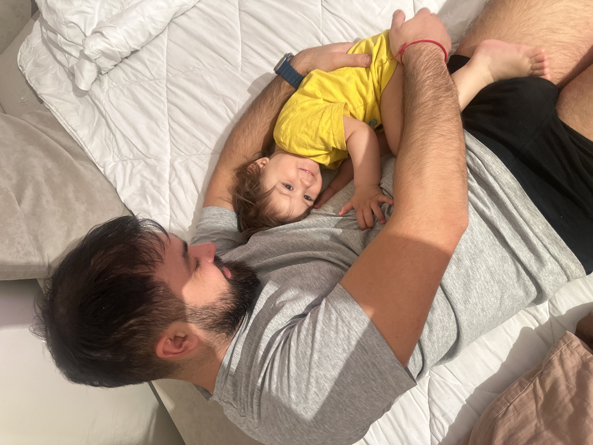 A man with dark hair and a beard is lying on a white comforter, cradling a toddler in a yellow shirt. The toddler is looking at the camera. The man is wearing a grey t-shirt and black shorts. The scene is indoors, likely a bedroom. There is a pillow to the left and possibly another to the right. The location is not evident from the image and cannot be determined to be Gavrilkovo, Russia.
