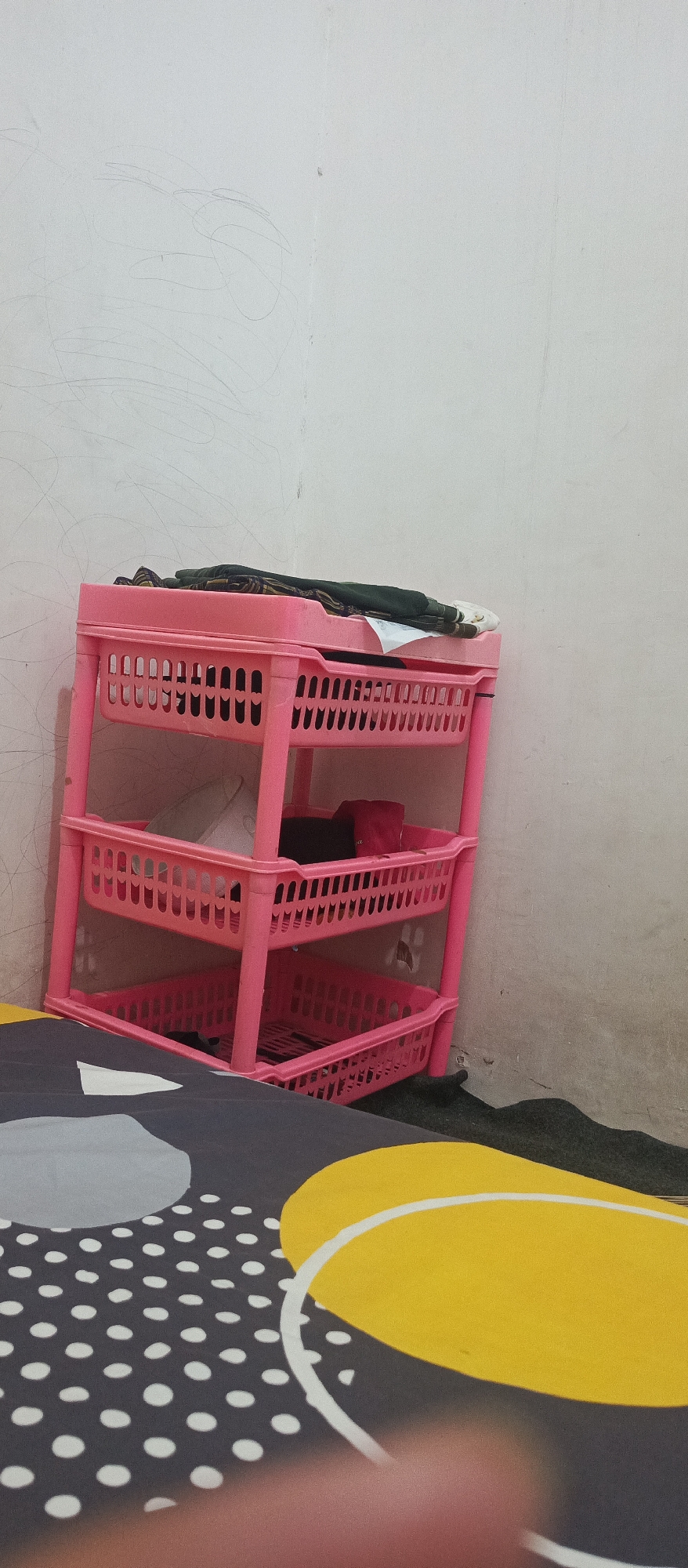 A pink three-tiered plastic shelf unit stands in the corner of a room, against a white wall with faint scribbled marks. The shelves are filled with various items, including folded clothing on the top shelf and what appears to be household goods in the lower shelves. A portion of a bed with a patterned duvet featuring grey, white, and yellow abstract shapes is visible in the foreground. The environment appears to be indoors, likely a bedroom or a storage area.