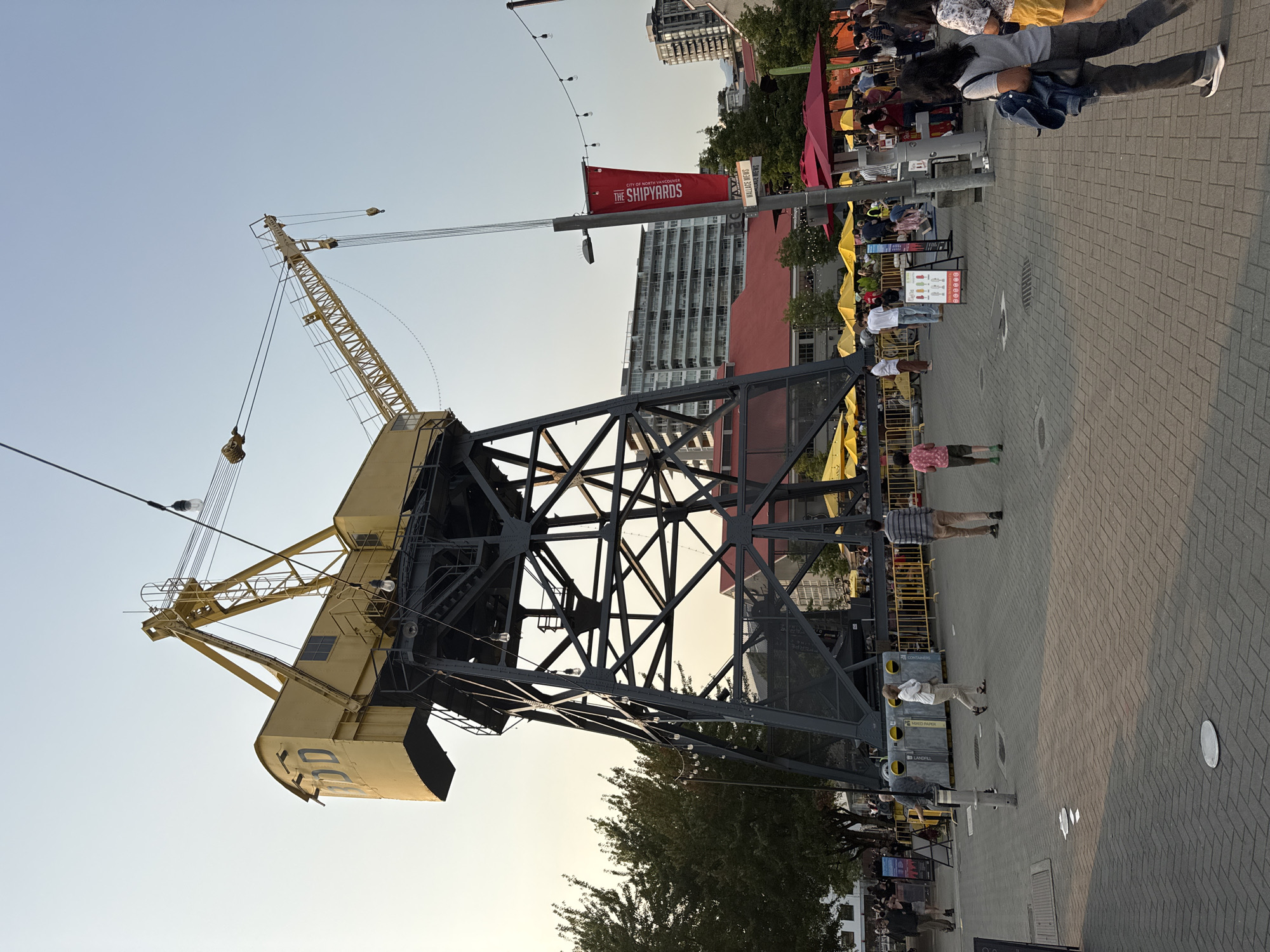 This image captures an outdoor scene at The Shipyards in North Vancouver, Canada. The focal point is a large, yellow industrial crane, its steel support structure dark gray. The crane's main housing is yellow with the letters "DDD" visible on its side. The crane is positioned in an open paved area where several people are present.
The environment includes a backdrop of modern buildings and trees. In the foreground, the paved ground is patterned with brickwork. There are yellow and red umbrellas shading outdoor seating areas, suggesting a public gathering space or outdoor dining. Several individuals are walking or standing, engaged in various activities. A red banner with white text "THE SHIPYARDS VANCOUVER" hangs from a pole. The sky is clear and light, indicating daytime. String lights are visible across the scene, some illuminated.