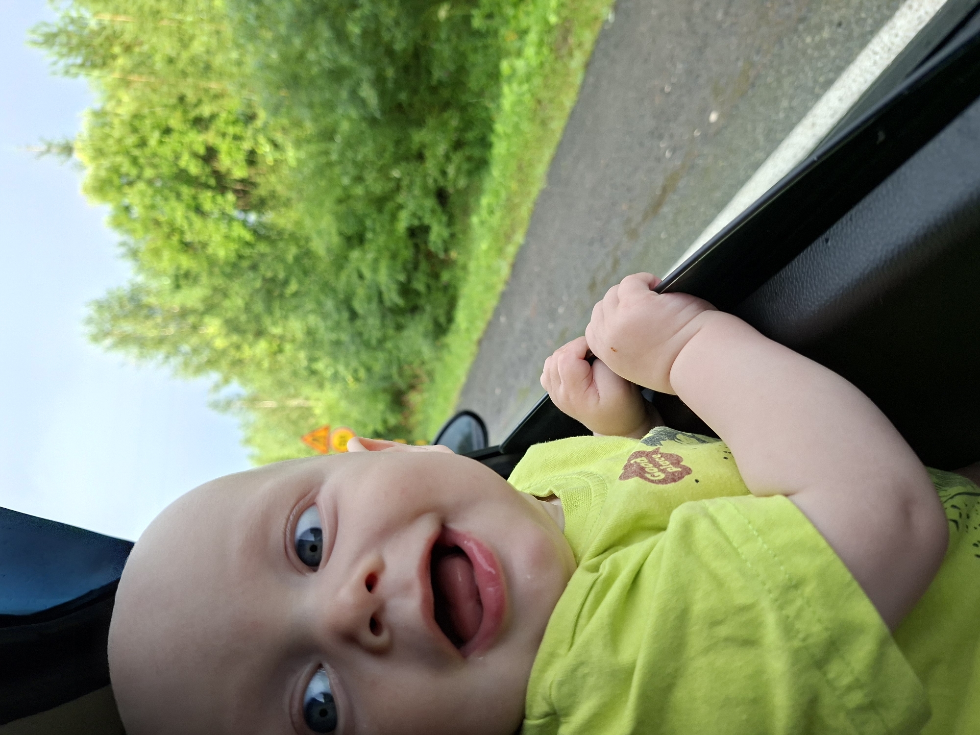A bald baby with wide blue eyes and an open mouth with a tongue visible is leaning out of a car window. The baby is wearing a bright green t-shirt with a circular graphic that says "Good place". The baby's hands are gripping the edge of the car door. In the background, there is a paved road and lush green trees. A yellow traffic sign is visible on the side of the road. The sky is overcast. This image was taken in the city of Yugo-Kamskiy, Russia.