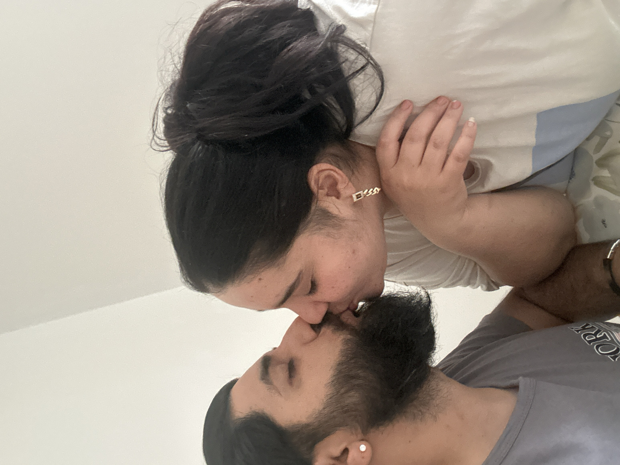 The image shows a close-up of a man and a woman kissing. The man has a beard and is wearing a gray t-shirt. The woman has her hair in a ponytail and is wearing a white shirt. They are both looking at each other with their eyes closed. The background is a plain white wall. There are no identifying markers or information to suggest this image was taken in Mainburg, Germany.
