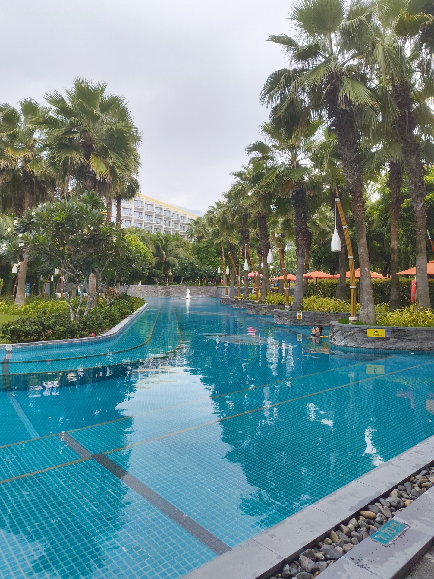 A long, winding swimming pool with turquoise blue tiles and darker grey borders is the central feature of this outdoor scene. The pool's surface reflects the overcast sky and the surrounding tropical vegetation. Palm trees with lush green fronds line the pool, casting shadows on the water. In the background, a large, multi-story hotel with a light yellow facade and white balconies is visible amongst the trees. Orange umbrellas are dotted along the poolside, suggesting an area for lounging. A single person is partially submerged in the water, appearing to be relaxing. A white sculpture, possibly a swan, is located in the distance within the pool. The edge of the pool is decorated with smooth, grey stones. This scene is located in the city of Phú Quốc, Vietnam.