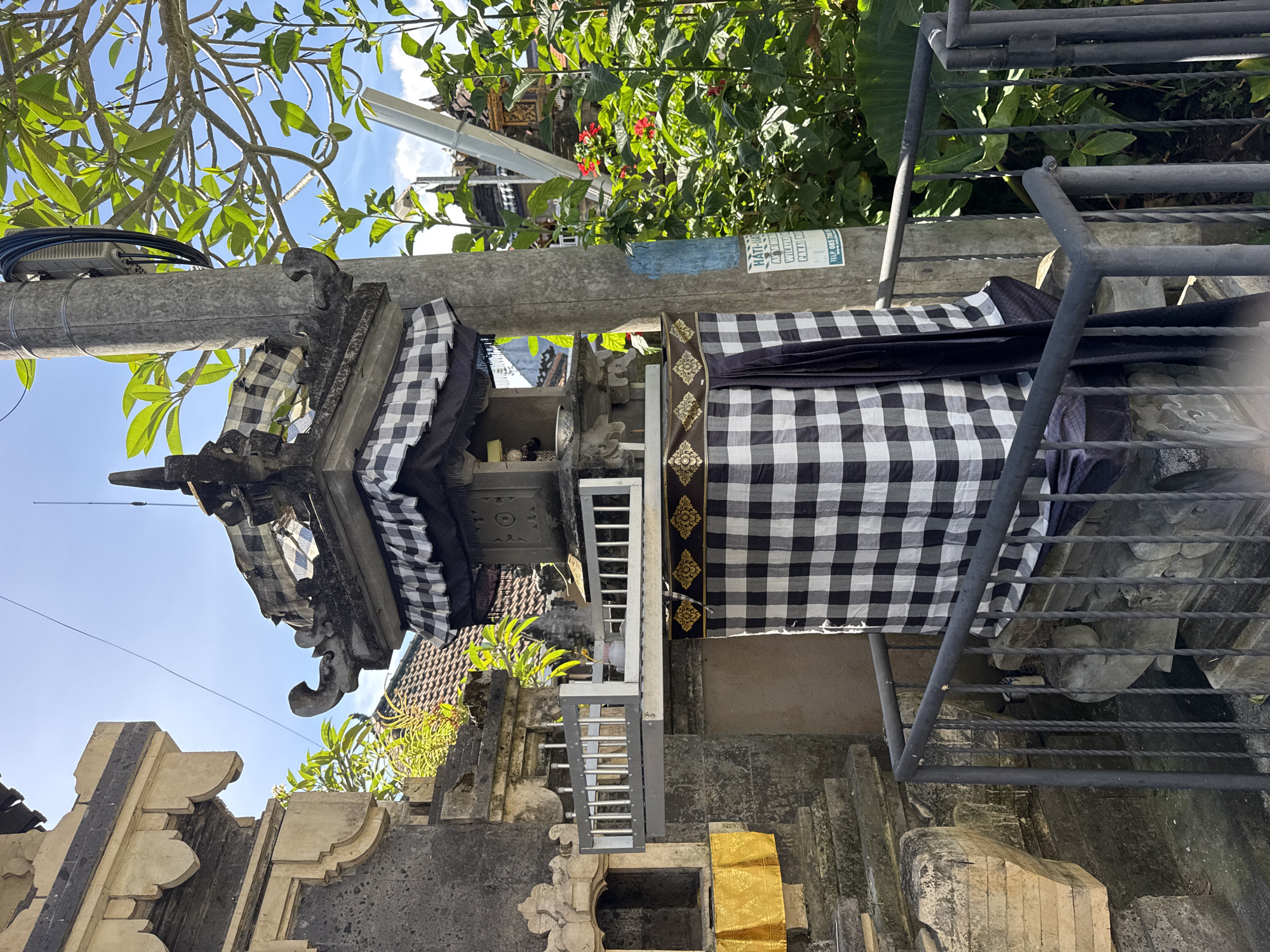 The image depicts a Balinese shrine or altar in Kuta Selatan, Indonesia. The shrine is adorned with a black and white checkered cloth and a gold-patterned sash. A metal staircase is located next to the shrine. The shrine is partially obscured by a utility pole and dense green vegetation. The sky is blue with some clouds visible in the background. The architecture of other buildings is partially visible in the background, indicating an urban or semi-urban setting.
